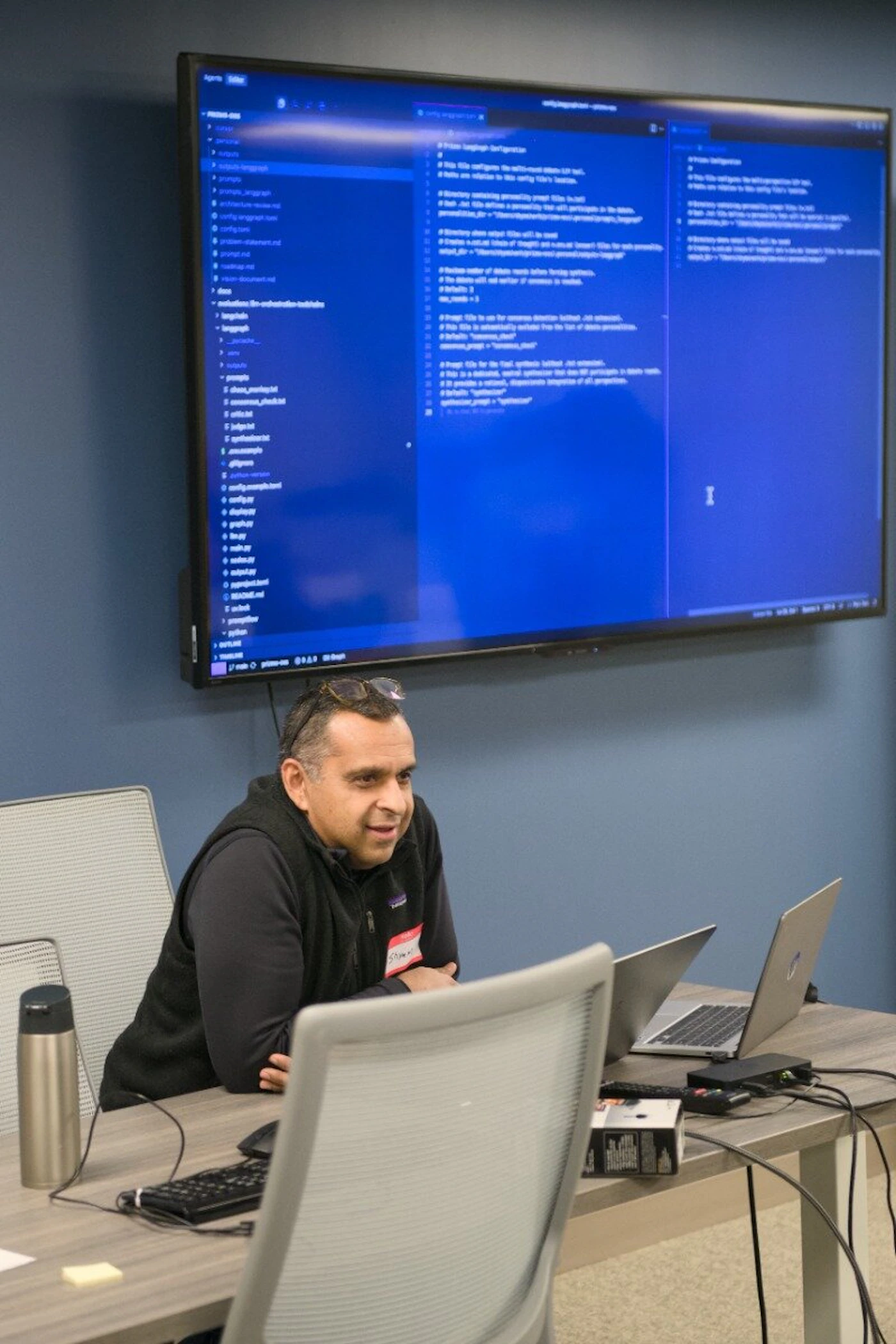 (Photo) A man sits at a conference table with laptops, positioned in front of a large wall-mounted monitor displaying software code. Text: PRISMS-OSS, personal, outputs, outputs-langgraph, prompts, prompts_langgraph, architecture-review.md, config.langgraph.yaml, config.yaml, problem-statement.md, prompt.md, roadmap.md, vision-document.md, docs, evaluations-llm-orchestration-toolchain, langchain, langgraph, main.py, README.md, Prisms-langgraph-Configuration, This file configures the multi-agent system., Directory containing personality prompt files (.txt), Number of return results before finishing synthesis., Prompt file to use for consensus detection (not-extension)., common_prompt = "consensus_check", Prompt file for the final synthesis (without .txt extension)., synthesizer_prompt = "synthesizer" person, office equipment, computer code | indoor office | large monitor with code, multiple laptops, name tag, water bottle | candid | Colors: #1e40af, #4b5563, #111827, #d1d5db Note: The image is a real-life photograph capturing a person in a professional office or meeting room setting.