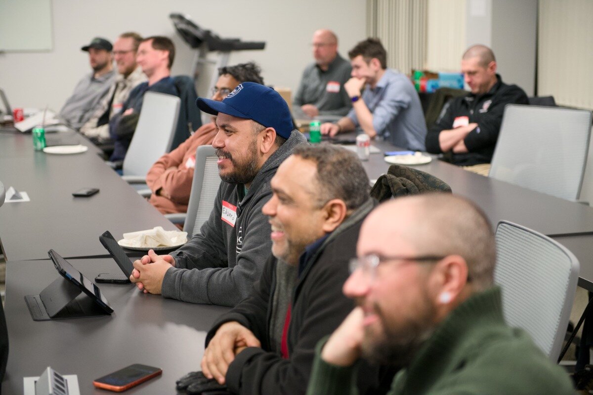 A group of men sitting at a long conference table in an office setting, appearing to listen to a presentation or participate in a meeting.. Text: Eduardo