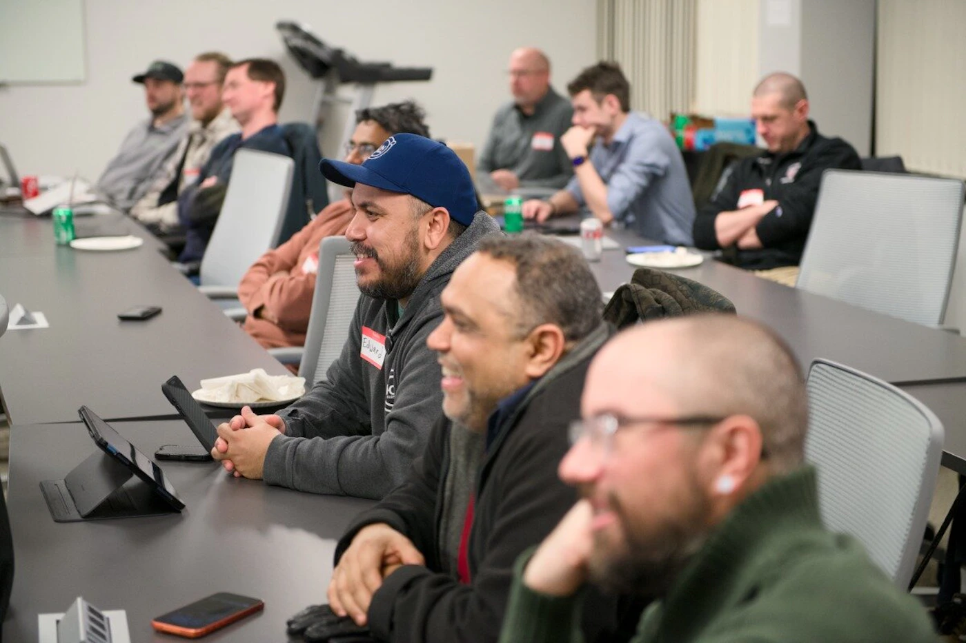 (Photo) A group of men sitting at a long conference table in an office setting, appearing to listen to a presentation or participate in a meeting. Text: Eduardo people in a meeting | indoor office | multiple people, conference table, name tags, laptops | candid | Colors: #333333, #D3D3D3, #1E3A8A, #4B5563 Note: The image is a real-life photograph capturing a group of people in a professional indoor environment.