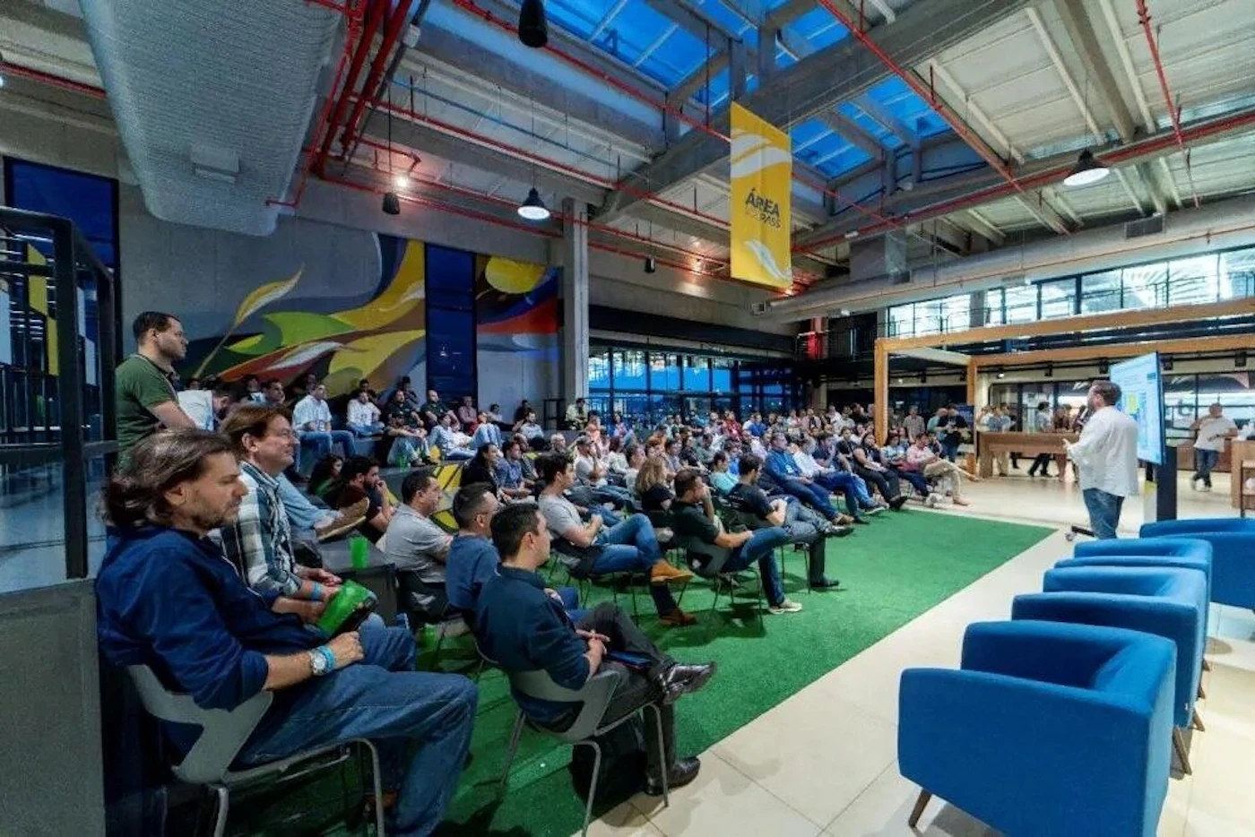 (Photo) A wide-angle photograph captures a presentation or conference event taking place indoors in a large, modern venue with industrial architecture and a high ceiling. A large audience is seated on chairs arranged on a green floor, listening to a male presenter standing near a vertical digital screen on the right. Text: ÁREA DE [PASSOS] Conference or business presentation with a large audience. | Indoor, large industrial/modern event space with exposed ductwork and a glass roof. | Large audience, abstract wall mural, yellow hanging banner, green floor covering, blue upholstered chairs in the foreground. | Candid/event photography, wide shot. | Colors: #4665A3, #788A8E, #C89F39, #004D00 Note: This is a captured image of a real-world event scene, characteristic of event photography.