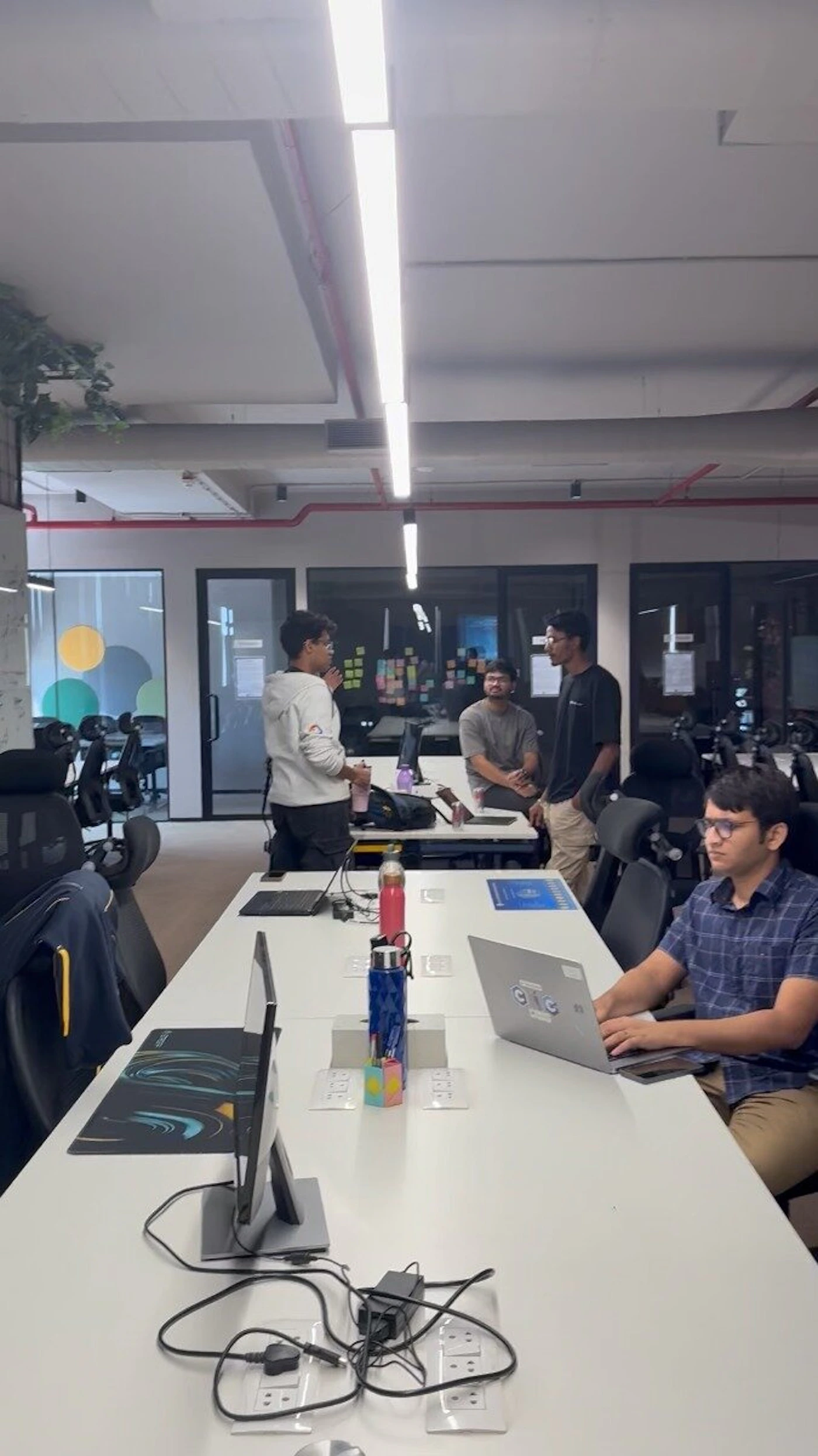 (Photo) A candid indoor shot of a modern office space where several young men are either working at a long white desk or engaged in a discussion in the background. Text: GDG people in an office | indoor | long white workstation, laptops, glass partitions, industrial-style ceiling with exposed pipes | candid | Colors: #FFFFFF, #2C2C2C, #7B7B7B, #000000 Note: The image is a real-life photograph capturing people and objects in a physical environment.