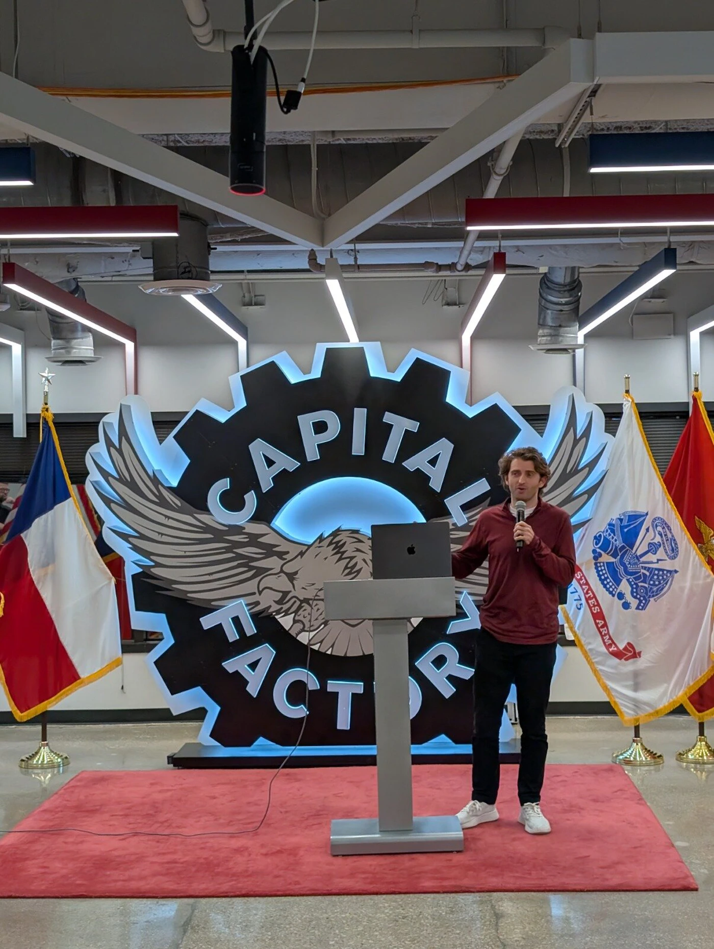 (Photo) A man wearing a maroon shirt and black pants is giving a presentation or speech at a modern podium, standing on a red carpet in an indoor event space. Text: CAPITAL FACTORY UNITED STATES ARMY 1775 person giving a presentation | indoor event space/auditorium | large branded backdrop (Capital Factory), flags (Texas flag, US Army flag), podium, red carpet | candid, event photography | Colors: #800000, #000000, #FFFFFF, #ADD8E6 Note: This image is a photograph capturing a real-world event, documenting a speaker giving a presentation in an indoor setting.