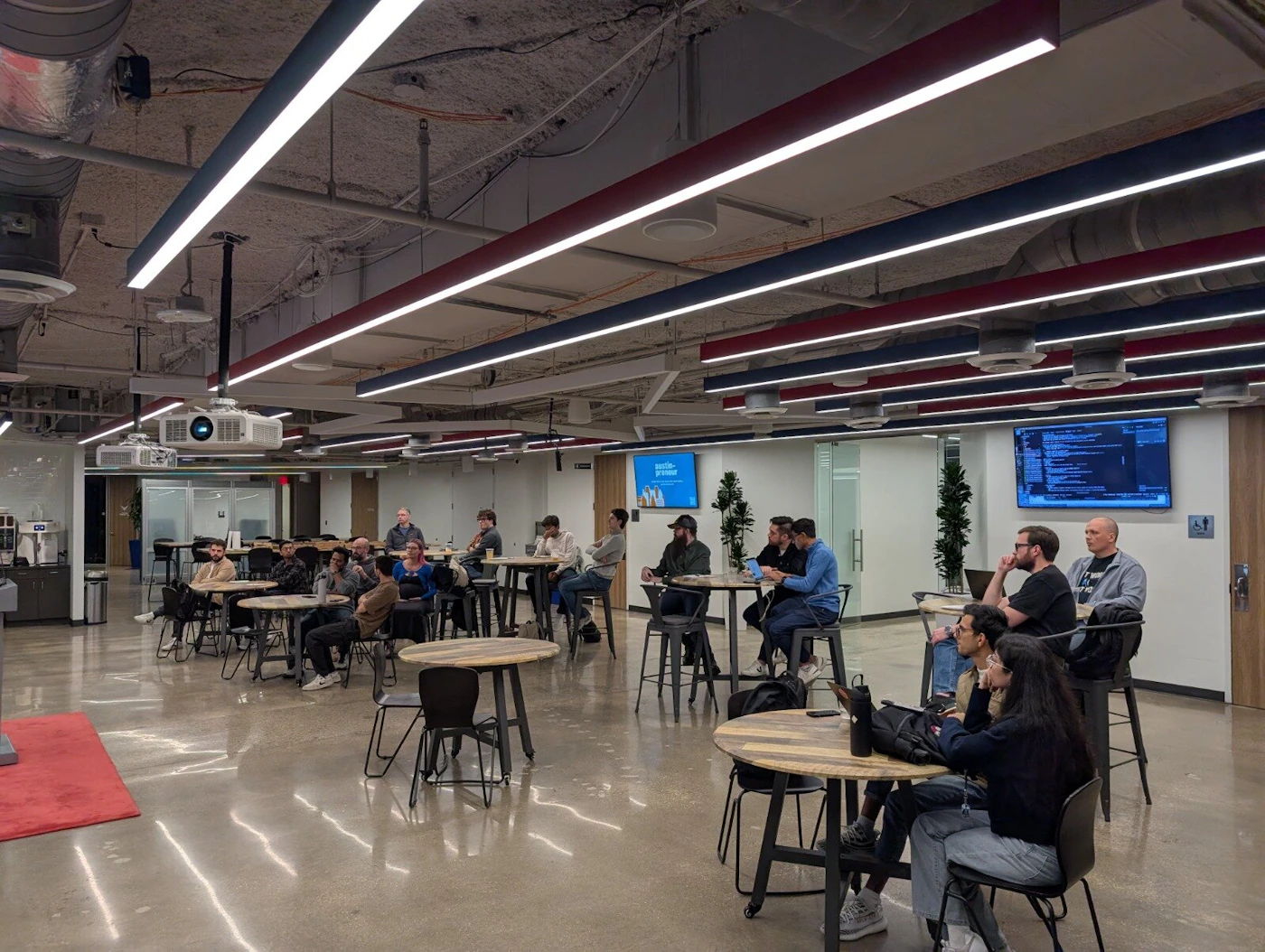 (Photo) A candid photograph captures a group of approximately 20 people seated at high-top tables in a large, modern office or communal space, gathered as if attending a presentation or informal meeting. Group of people attending an event/meeting | Indoor, modern corporate office or co-working space | Polished concrete floor, exposed ceiling structure, linear lighting fixtures with red and blue accents, projector, and multiple screens displaying code/slides. | Candid, wide shot | Colors: #404040, #C0C0C0, #A0522D, #FF0000, #000080 Note: The image is a digital photograph capturing a real-world indoor scene featuring people, office architecture, and technical equipment.