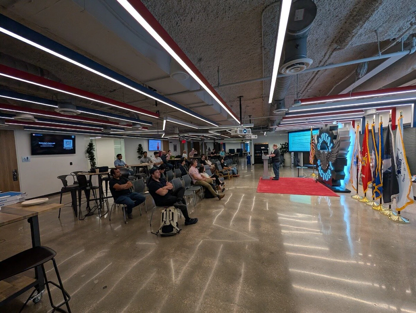 (Photo) A man delivers a presentation to a seated audience in a modern, industrial-style event space featuring a large 'Capital Factory' logo and a row of military flags. Text: CAPITAL FACTORY, UNITED STATES COAST GUARD presentation | indoor | audience, speaker, military flags, large gear-shaped logo sculpture, industrial ceiling with red and blue lighting | wide-angle candid | Colors: #a9a9a9, #e62020, #00ffff, #ffffff Note: The image is a real-world photograph capturing a live event in an indoor environment with natural lighting and depth of field.