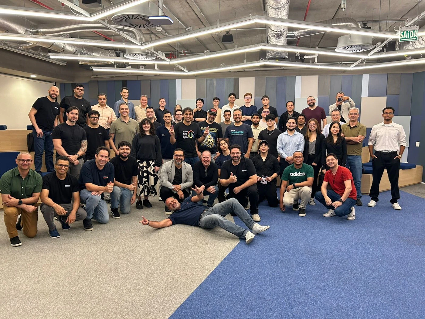 (Photo) A large group of approximately 48 people, likely colleagues, pose for a group photo inside a modern office space featuring exposed ceilings and blue accent walls. Text: SAÍDA adidas I AM DATA DRIVEN [partially obscured] Large group of professional colleagues/team | Indoor, modern office/meeting space with high ceilings and modular seating | Many people (around 48) arranged in standing and kneeling rows; one person lying playfully in the foreground. | Group portrait/candid | Colors: #6A6A6A, #374B8D, #000000, #E0E0E0 Note: The image is a captured moment of a large gathering of people posing for a picture in an indoor setting, characteristic of a group portrait photograph.