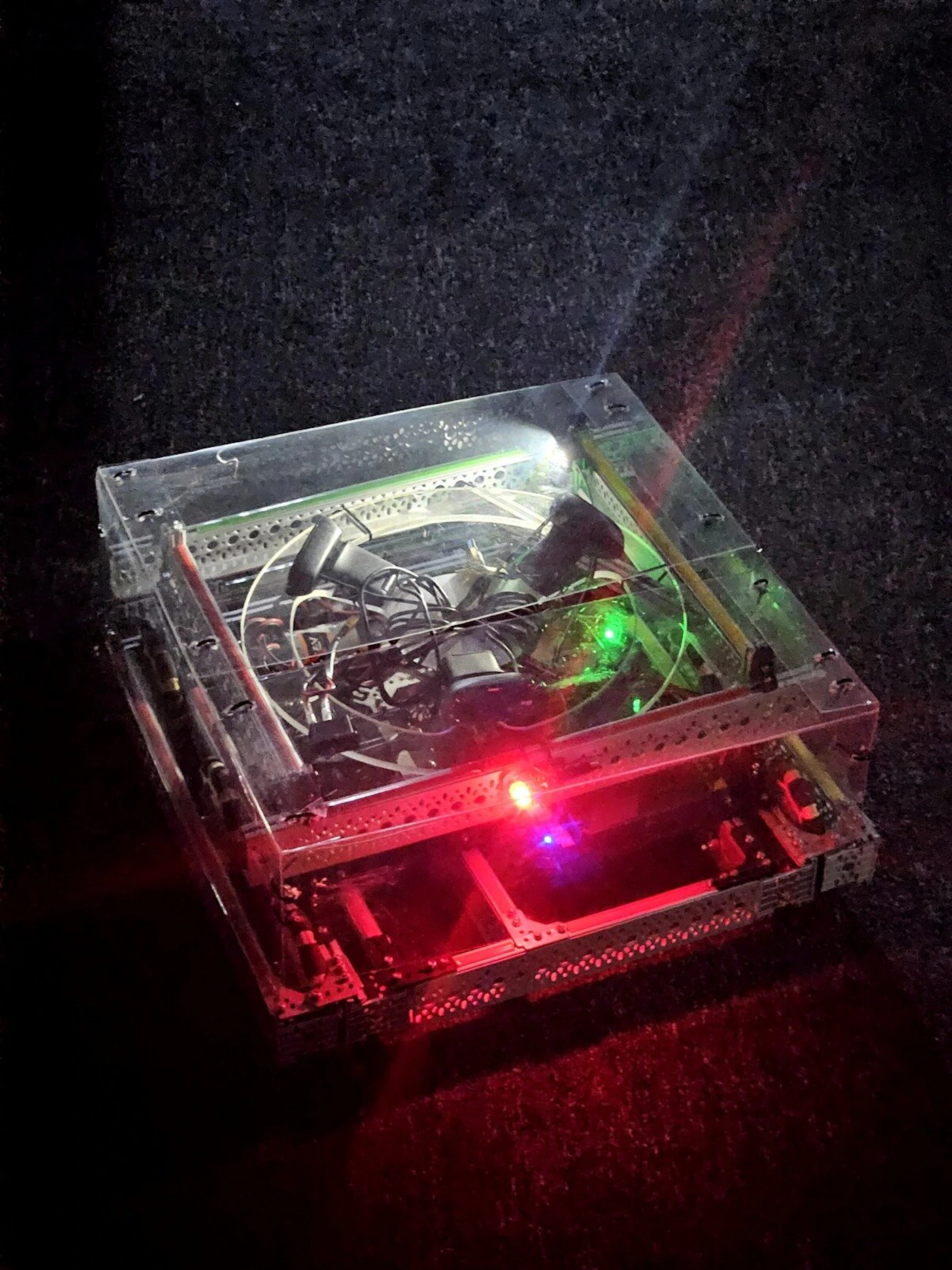 (Photo) A small, box-shaped robot with a transparent acrylic top and visible internal wiring is illuminated by glowing red and green lights on a dark, textured surface. robot | indoor | glowing LEDs, transparent casing, internal electronics, metal frame | low-light photography | Colors: #000000, #FF0000, #00FF00, #C0C0C0 Note: The image is a real-world photograph capturing a physical object, specifically a custom-built robot, in a low-light environment.