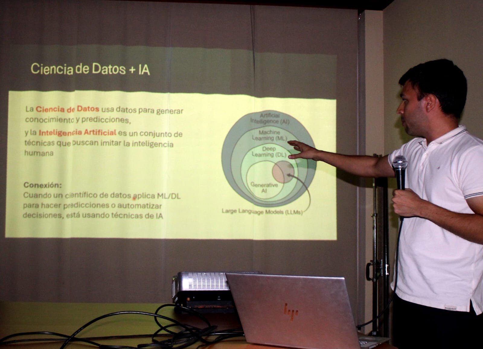(Photo) A man in a white polo shirt is giving a technical presentation on Data Science and AI, pointing toward a projected slide that contains Spanish text and a concentric circle diagram of AI concepts. A laptop and a projector are visible on the table in the foreground. Text: Ciencia de Datos + IA La Ciencia de Datos usa datos para generar conocimiento y predicciones, y la Inteligencia Artificial es un conjunto de técnicas que buscan imitar la inteligencia humana Conexión: Cuando un científico de datos aplica ML/DL para hacer predicciones o automatizar decisiones, está usando técnicas de IA Artificial Intelligence (AI) Machine Learning (ML) Deep Learning (DL) Generative Al Large Language Models (LLMs) person giving a presentation | indoor (conference room/classroom) | projected slide, technical diagram, microphone, laptop, projector | candid/event photography | Colors: #FFFFFF, #000000, #A9A976, #4A4A4A Note: The image is a candid photograph capturing a real-world event—a speaker giving a presentation in an indoor setting.