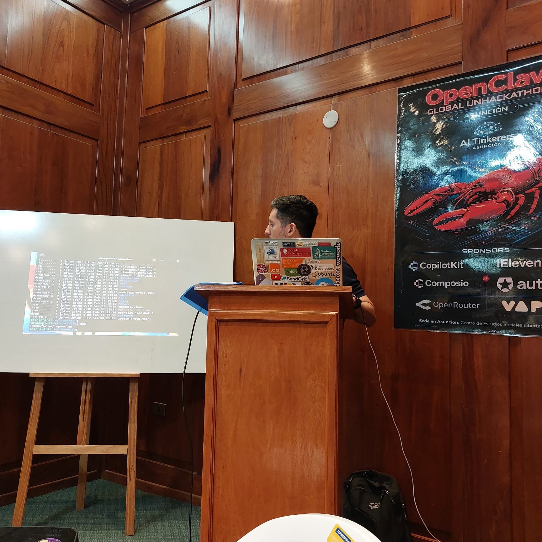 (Photo) A man stands behind a wooden podium giving a presentation, with a terminal window projected onto a whiteboard and an event poster for a hackathon on the wood-paneled wall. Text: OpenClaw GLOBAL UNHACKATHON ASUNCIÓN AI Tinkerers ASUNCIÓN SPONSORS CopilotKit Composio OpenRouter ElevenLabs auth... VAP Sede en Asunción: Centro de Estudios Sociales Libertad jpino jpino clawbot.json openclaw.json update_check.json Google SendGrid Ubuntu wework person | indoor | wooden podium, whiteboard with projection, event poster, wood-paneled walls | candid | Colors: #8b4513, #ffffff, #000000, #c0c0c0 Note: This is a real-life photograph capturing a person, a podium, and a whiteboard in an indoor setting.