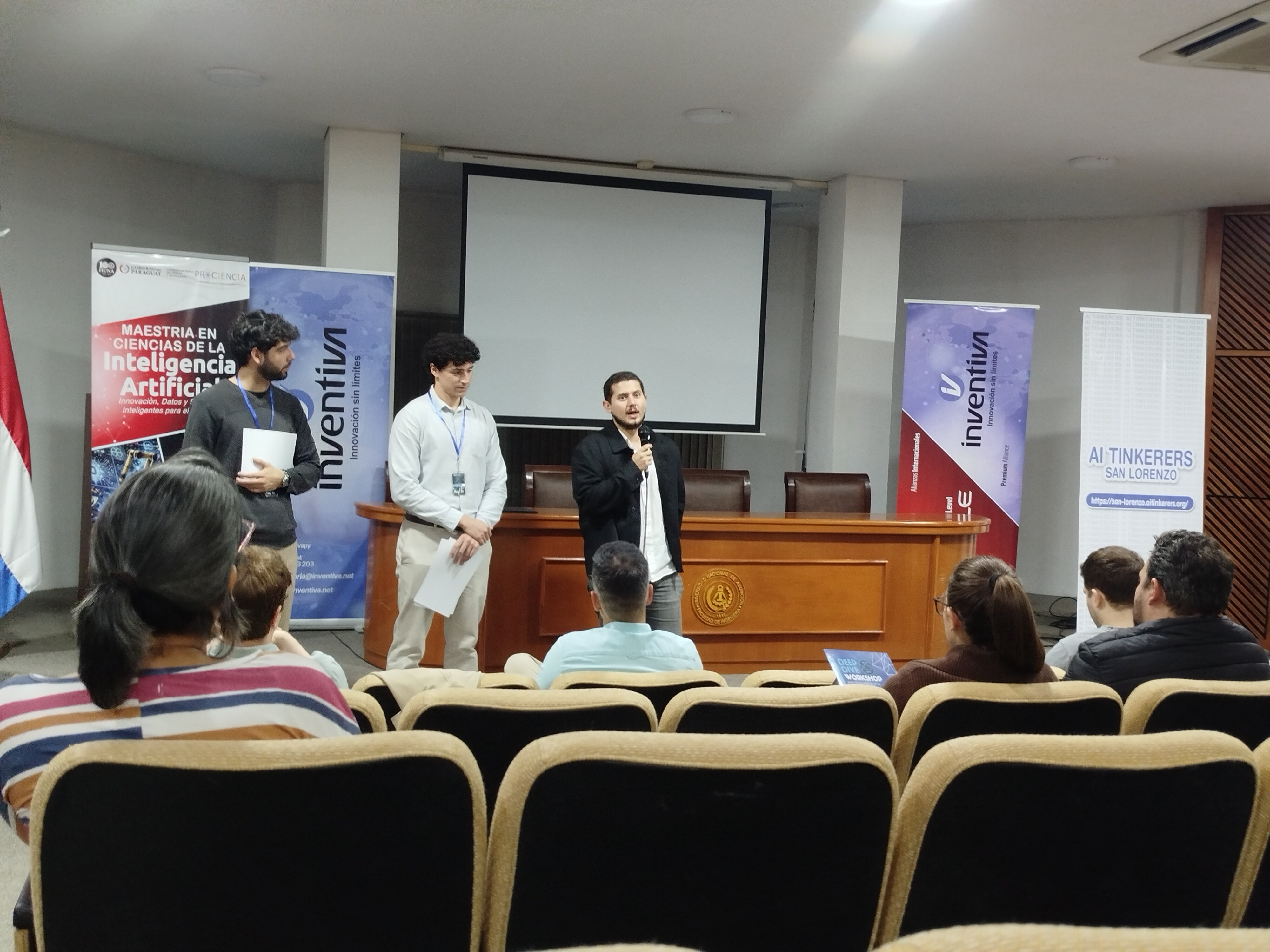 (Photo) A man is giving a presentation with a microphone in front of an audience in an auditorium, flanked by two other men and several promotional banners. Text: MAESTRIA EN CIENCIAS DE LA Inteligencia Artificial, inventiva Innovación sin límites, AI TINKERERS SAN LORENZO, https://san-lorenzo.ai-tinkerers.org/ Presentation or academic event | Indoor auditorium or lecture hall | Speakers, audience, promotional banners, projector screen | Candid event photography | Colors: #FFFFFF, #8B4513, #2E3192, #ED1C24 Note: This is a real-life photograph capturing a live event with people in an indoor setting.
