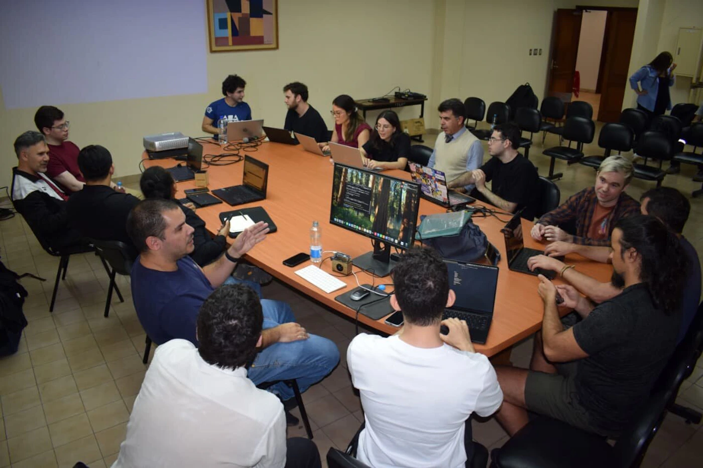 (Photo) A group of people sitting around a large wooden conference table filled with laptops, collaborating and discussing in a meeting room. people | indoor | group around a conference table with laptops | candid | Colors: #D2B48C, #2E3B4E, #FFFFFF Note: The image is a real-life photograph capturing a group meeting indoors.