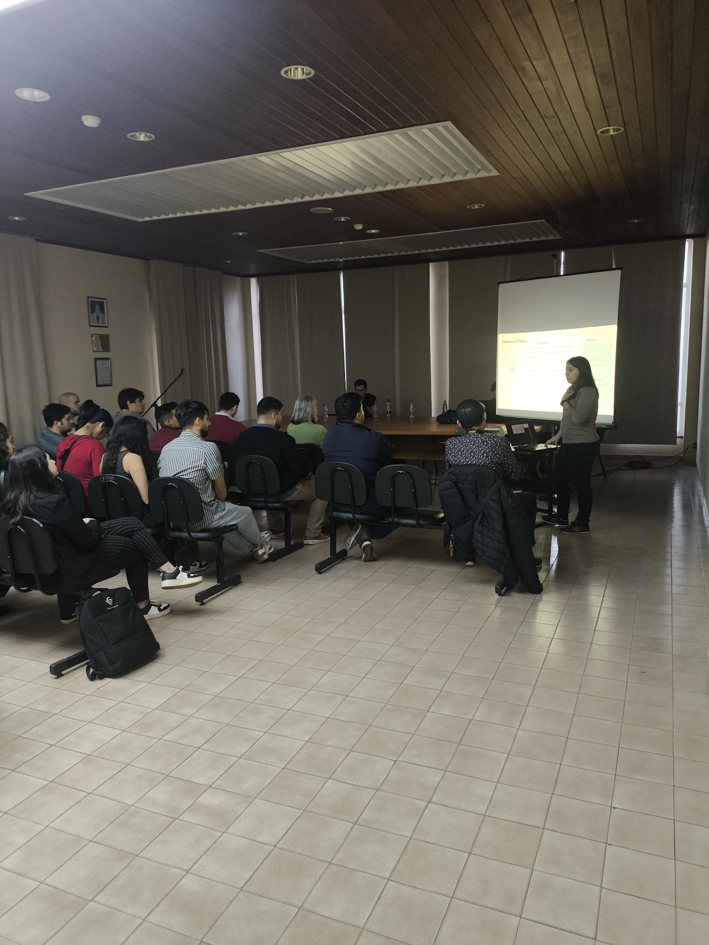 (Photo) A photograph captures an indoor presentation or lecture where a woman stands next to a projection screen addressing an audience seated in rows of chairs. The room features a dark wood-paneled ceiling and light tiled floors. Text: Differentiation Points, Format, Styles, [unreadable] Group of people attending a presentation | indoor (conference room or lecture hall) | Presentation screen, audience seating, wood ceiling paneling | Candid, documentary | Colors: #6F4F3B, #C8C0B4, #000000 Note: This is a candid photograph documenting a real-world event (a presentation) taking place indoors.