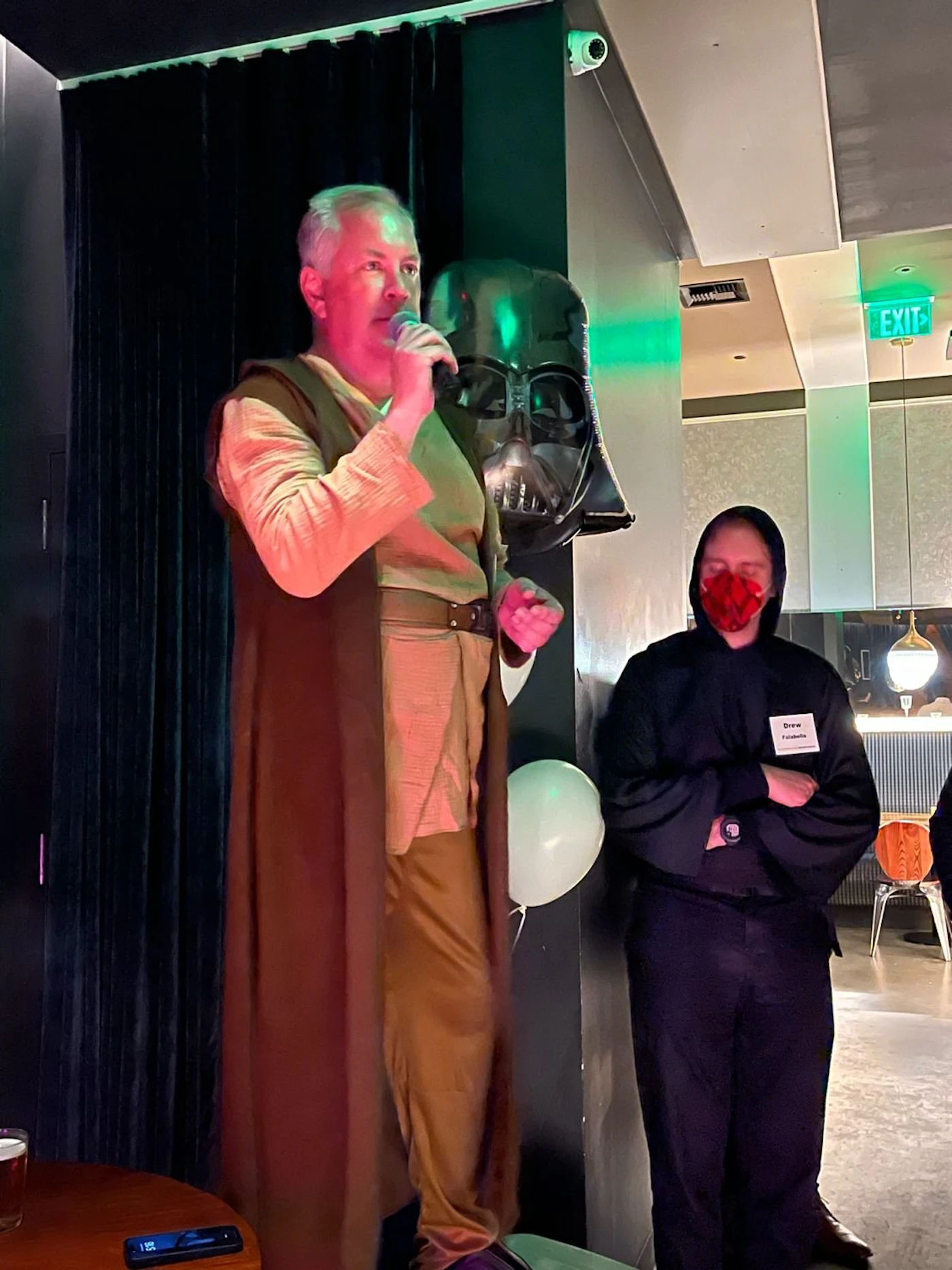 (Photo) A man dressed in a Jedi costume speaks into a microphone on a stage, with a Darth Vader balloon behind him and a person wearing a name tag standing nearby. Text: Drew people | indoor | costumes, microphone, balloon, event setting | candid Note: This is a real-life photograph of a costumed person speaking at an event, captured with a camera.