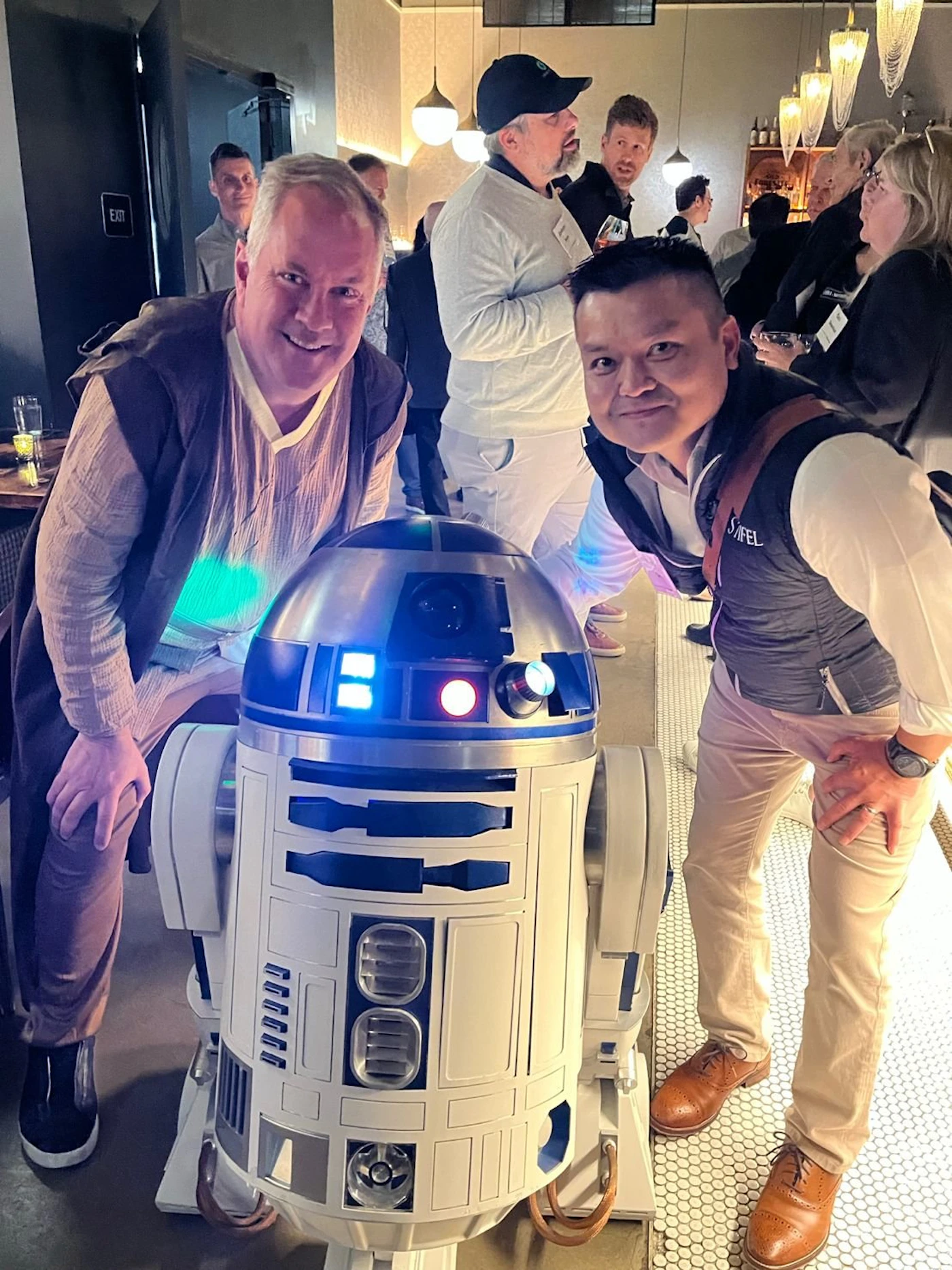 (Photo) Two men pose with an R2-D2 droid from Star Wars in what appears to be a bar or restaurant. Text: EXIT people, object | indoor | Star Wars droid, social gathering | candid Note: The image is a real-life photograph of people and an object in a social setting.