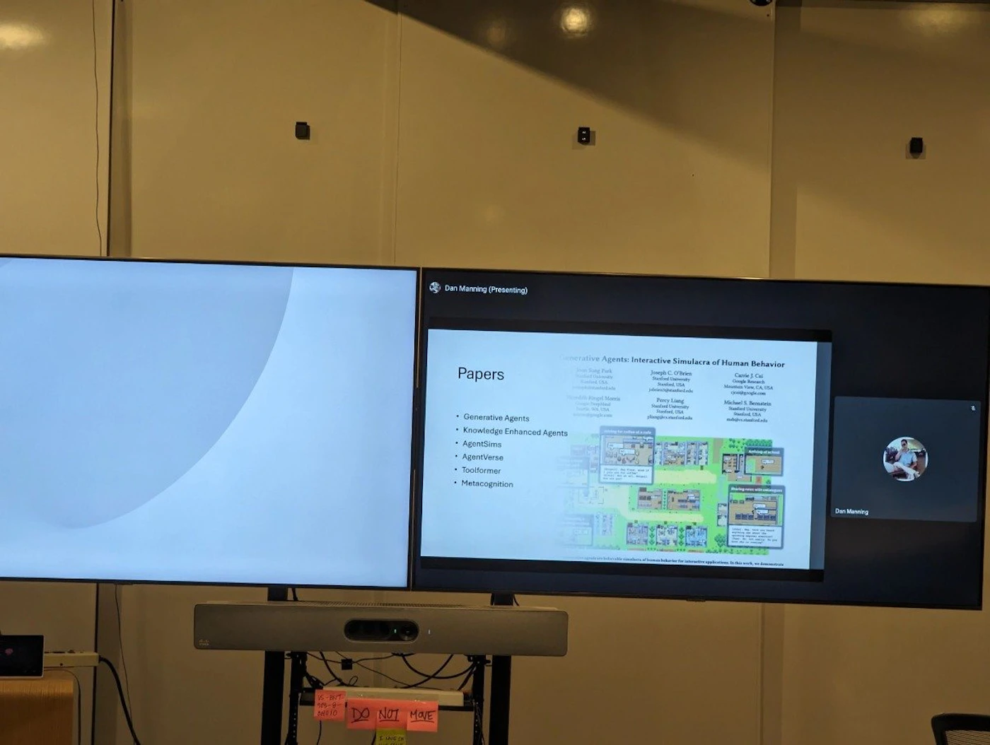 (Photo) A photo of a presentation being given on a large screen, focusing on generative agents and interactive simulacra of human behavior. A smaller screen shows the presenter and a list of papers. Text: Dan Manning (Presenting) Papers Generative Agents Knowledge Enhanced Agents AgentSims AgentVerse Toolformer Metacognition 8 Generative Agents: Interactive Simulacra of Human Behavior DO NOT MOVE presentation screen | indoor | presentation slides, presenter video, multiple screens | informal documentation Note: The image is a real-world photograph of a presentation taking place in a room. It captures the environment, screens, and presentation content.