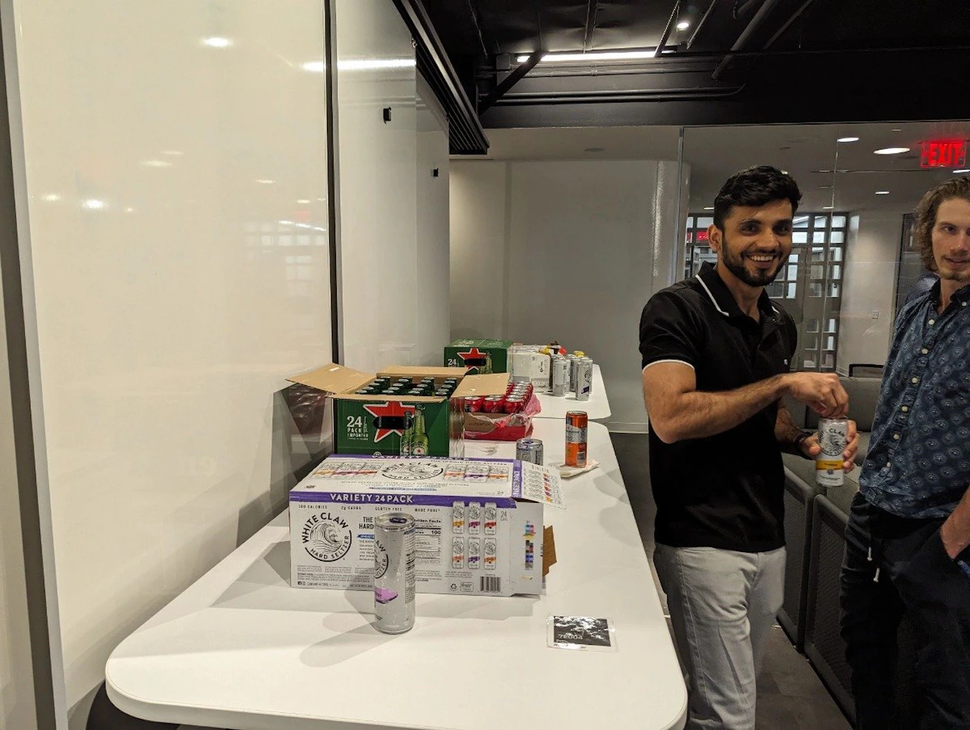(Photo) A photo of two people standing near a table with drinks and boxes. One person is opening a can. Text: 24 PACK VARIETY 24 PACK people | indoor | multiple people, drinks, table | candid Note: The image is a real-life captured scene of people and objects in an indoor environment, making it a photograph.