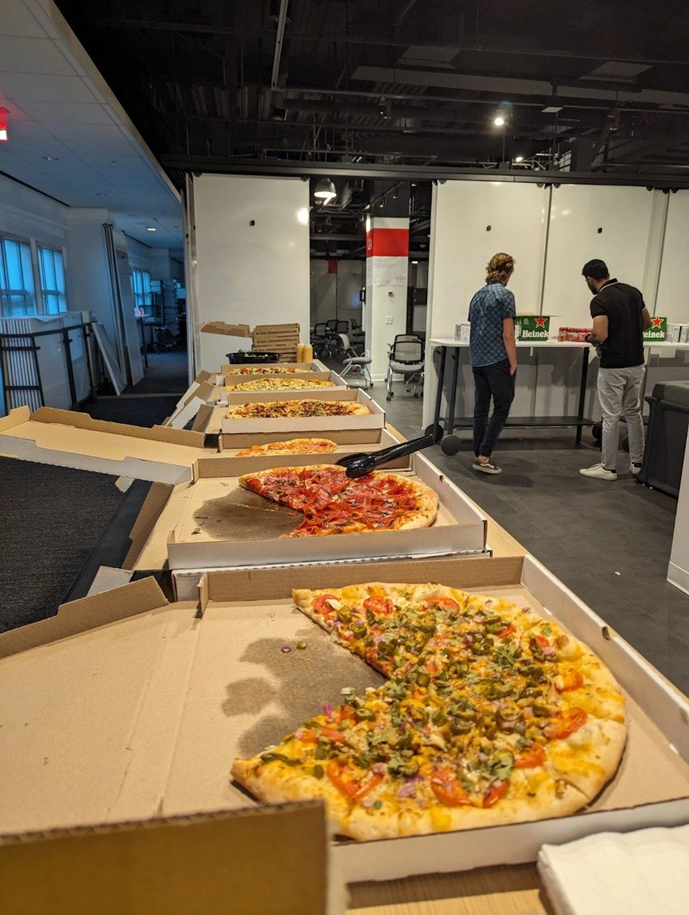 (Photo) A photo of several pizza boxes open on a table, with some slices missing. Two people are standing in the background near a counter. Text: Heinek food, people | indoor | multiple pizzas, partially eaten, people in background | candid Note: The image is a real-life captured image of a scene, rather than a drawing, logo, or banner.  It depicts a moment in time.