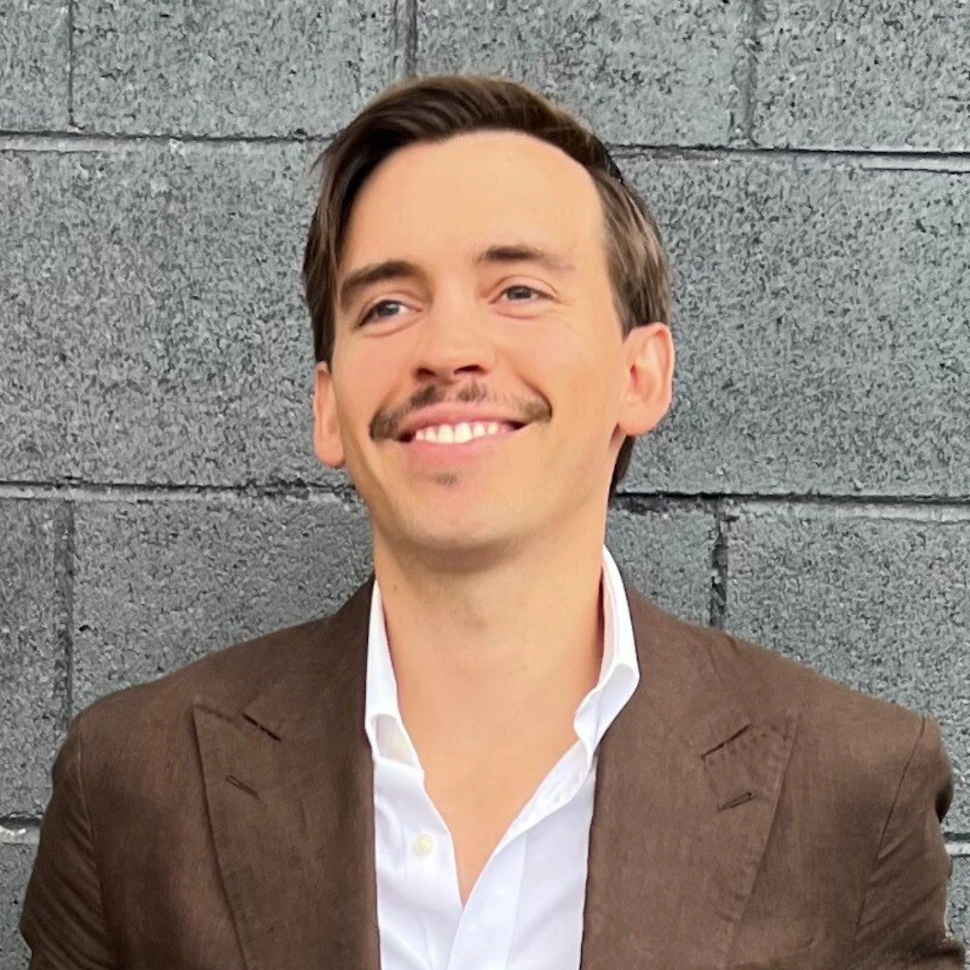 (Photo) A headshot of a smiling man with a prominent mustache, wearing a brown suit jacket over a white collared shirt, against a dark gray, textured brick wall. person (man with a mustache) | outdoor/urban setting against a textured gray wall | smiling subject, brown jacket, white shirt | portrait/headshot | Colors: #704332, #FFFFFF, #666666 Note: The image is a realistic capture of a person, characteristic of a photograph or headshot.