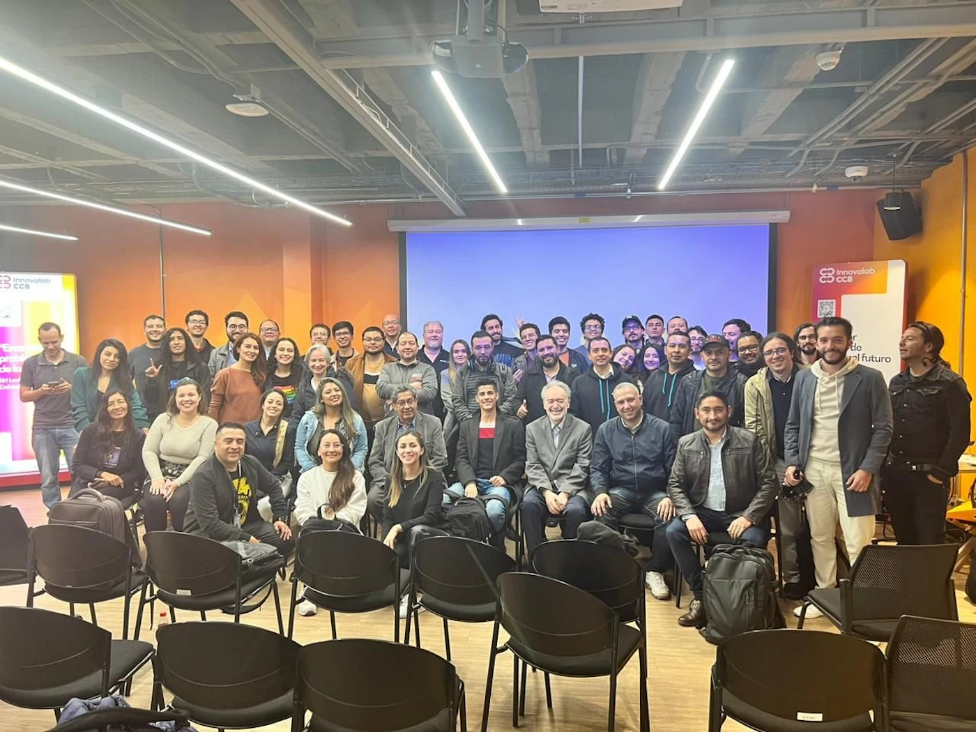 (Photo) A large group of people poses for a photo in a presentation room.  They are standing and sitting in front of empty chairs and a large screen. people | indoor | large group, presentation room | group photo Note: The image is a real-life captured image of a group of people, clearly a photograph of an event.  It's not a logo, illustration, banner, or handwriting.