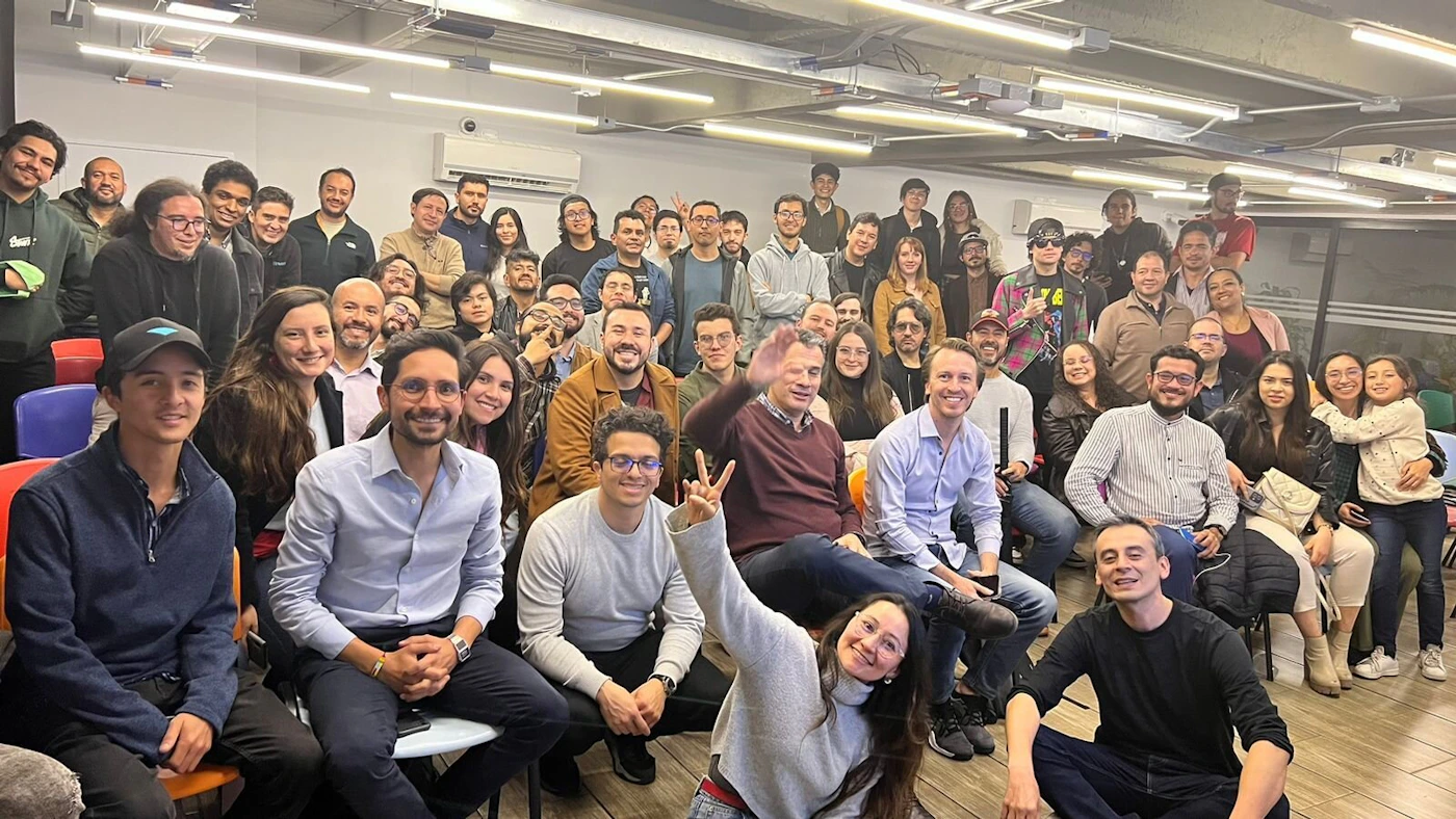 (Photo) A large group of people are gathered together, possibly for a meeting or social event. They are sitting and standing in rows, smiling and posing for the camera. people | indoor | large group, casual attire | candid Note: The image is a real-life captured moment of a group of people, making it a photograph.  It doesn't contain any designed elements suggesting other categories like a logo or banner.