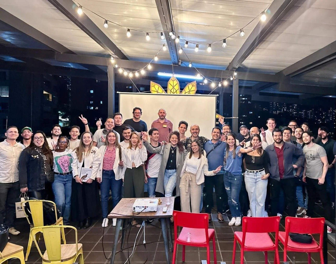(Photo) A group of people are posing for a photo, some making hand gestures, in what appears to be an outdoor or rooftop setting with string lights and a projector screen behind them. people | outdoor | group photo, string lights, projector screen | candid Note: The image is a real-life captured moment of a group of people, indicating a photograph. It's not a logo, illustration, or other specific type.