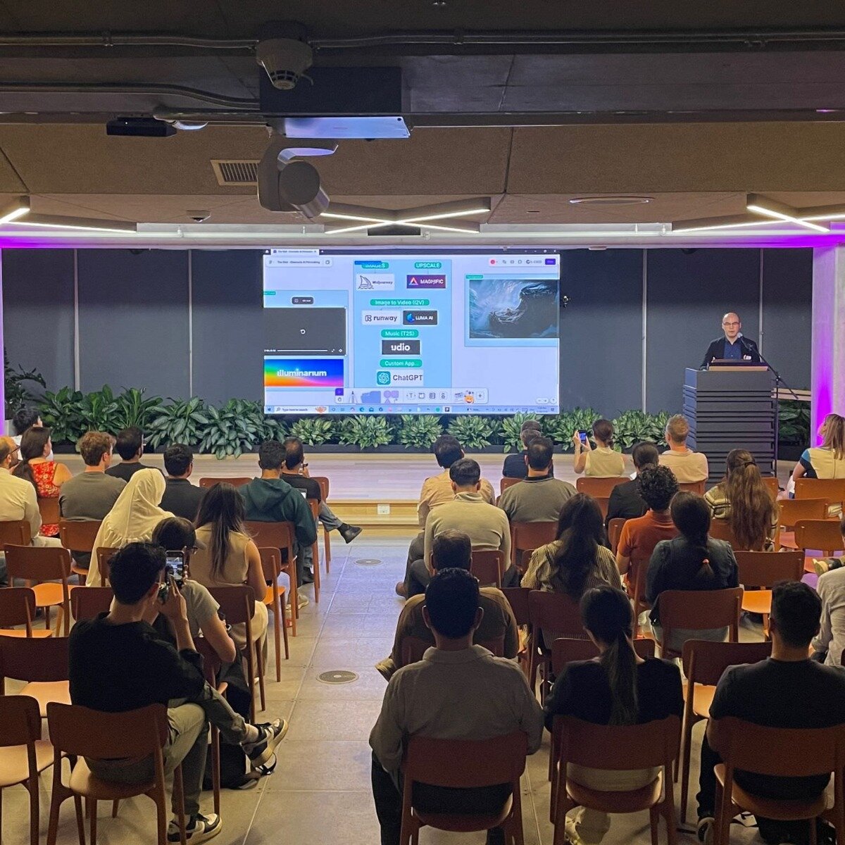 (Photo) The image captures an indoor event with an audience seated in rows facing a large screen and a presenter at a podium. The screen displays various software interfaces and names related to AI and creative tools. Text: Image to Video (I2V) Runway Luma.AI Music (T2A) udio Custom App ChatGPT Illuminarium UP SCALE WASHPIO presentation, conference, audience, technology | indoor, auditorium or large conference room | a speaker at a podium, a large screen displaying software applications, an attentive audience, ambient purple lighting | candid, event photography | Colors: #8B4513, #4CAF50, #808080, #FFFFFF, #8A2BE2 Note: This image is a direct capture of a real-world scene, featuring people, a room, and a presentation setup, which is characteristic of a photograph.