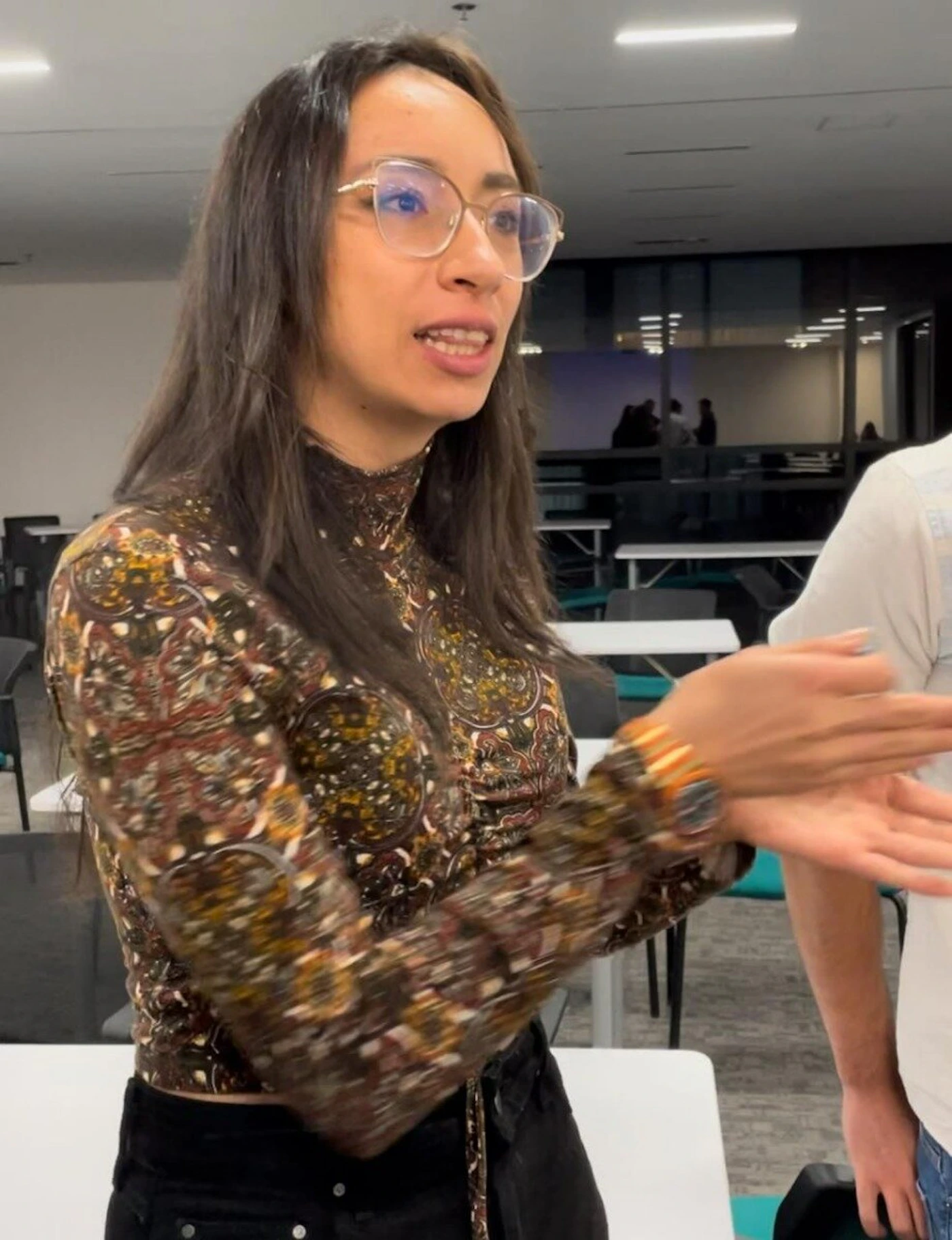 (Photo) A woman with glasses and a patterned long-sleeved shirt is gesturing with her hands, seemingly in conversation, in what appears to be an office or common area. person | indoor | gesturing, casual conversation | candid Note: The image is a real-life captured moment, depicting a person in a casual setting.  It exhibits characteristics of a candid photograph.