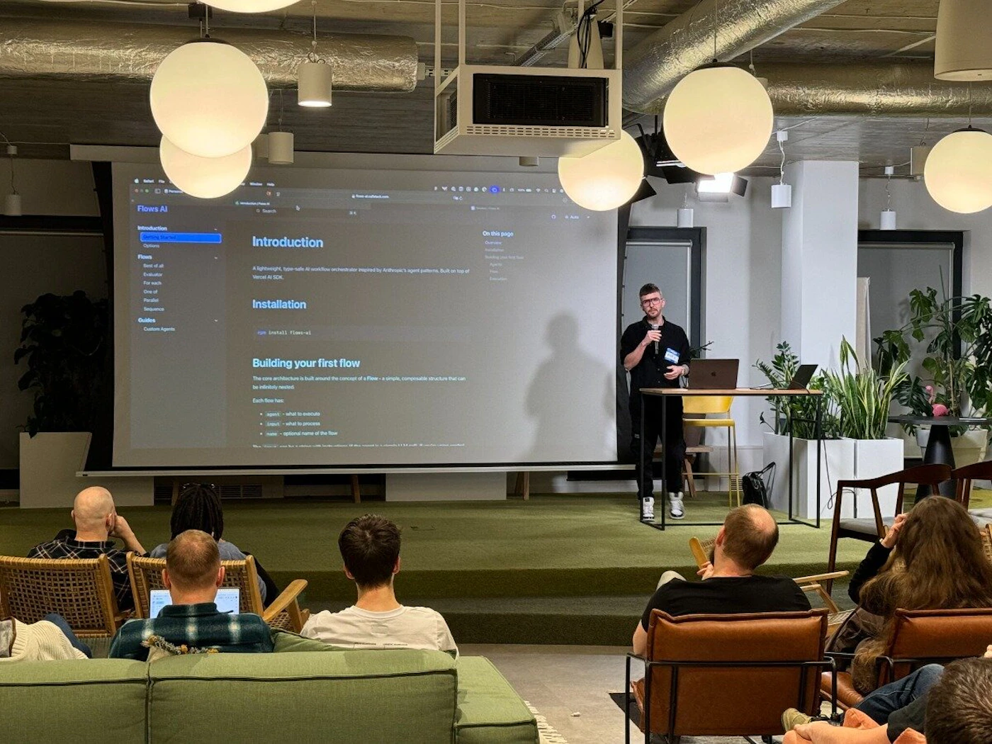 (Photo) A presenter stands in front of a projected screen displaying code and documentation, addressing an audience seated in chairs and on a couch. Text: Flows AI Introduction A lightweight, type-safe rails for workflow orchestration powered by agent patterns. Built on top of Ray. Installation pip install flows-ai Building your first flow The entire architecture is built around the concept of a Flow - a piece, composable resource that can execute arbitrary functions. Each Flow consists of: - agents - what is executed - inputs - arguments registered to the Flow - outputs - return values of the Flow people | indoor | presentation, audience, projector screen | candid Note: The image is a real-life captured scene of a presentation, making it a photograph. It's not a logo, banner, illustration, or handwriting.