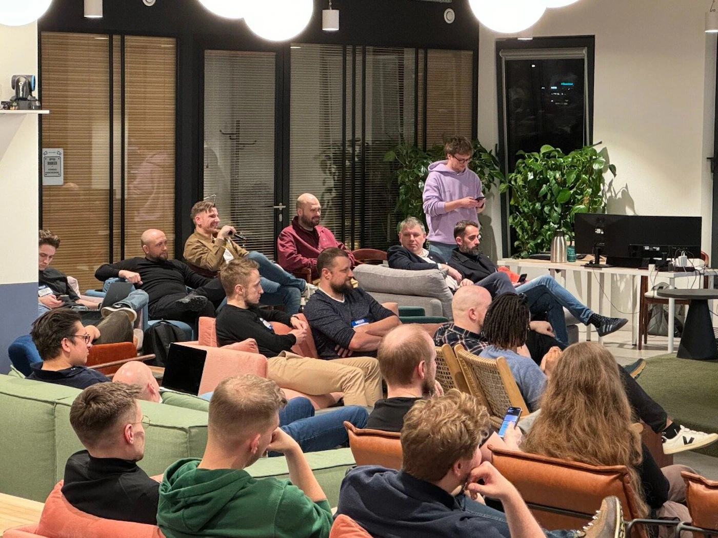 (Photo) A group of people are sitting on couches in a casual office setting, some looking at a presentation on a screen, others looking at their phones. people | indoor | group of people, office setting, presentation | candid Note: The image is a realistic depiction of a scene, captured with a camera, making it a photograph. It's not a drawing, logo, or other graphic type.