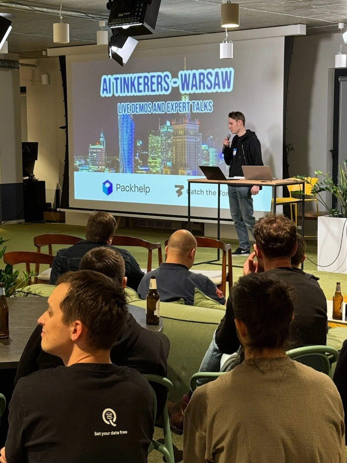 (Photo) A speaker presents to an audience in a room with a projector screen displaying the event title and sponsors. The audience is seated and listening to the presentation. Text: AI TINKERERS - WARSAW LIVE DEMOS AND EXPERT TALKS Packhelp Catch the Ton Set your data free people | indoor | presentation, audience, projector screen | candid Note: The image captures a real-life event with people and a presentation, making it a photograph. It's not a staged advertisement or illustration.