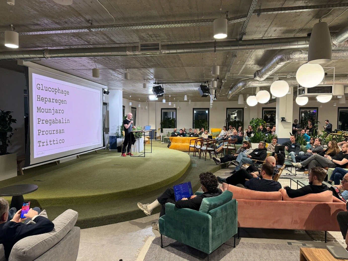 (Photo) A photo of a person presenting to a seated audience in a modern office space. The presenter stands on a small stage in front of a large screen displaying a list of words. Text: Glucophage Heparegen Mounjaro Pregabalin Proursan Trittico people | indoor | presentation, audience, modern office | candid Note: The image is a real-life captured scene of a presentation, depicting people and a venue. This makes it a photograph.