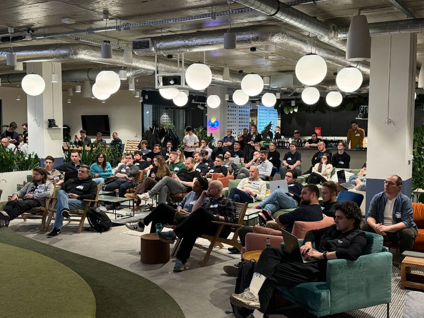 (Photo) A photo of a group of people sitting in a modern, open-plan office space, appearing to be attending a presentation or meeting. people | indoor | group of people, presentation setting, office environment | candid Note: The image is a real-life captured image of a scene with people in an office setting.  It exhibits characteristics of a candid photograph.