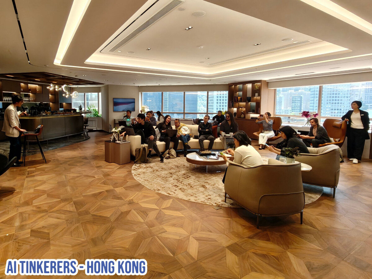 The image shows a group of people gathered in a modern office or event space, possibly attending a presentation or meeting. There is a speaker standing in front of the group.. Text: AI TINKERERS - HONG KONG