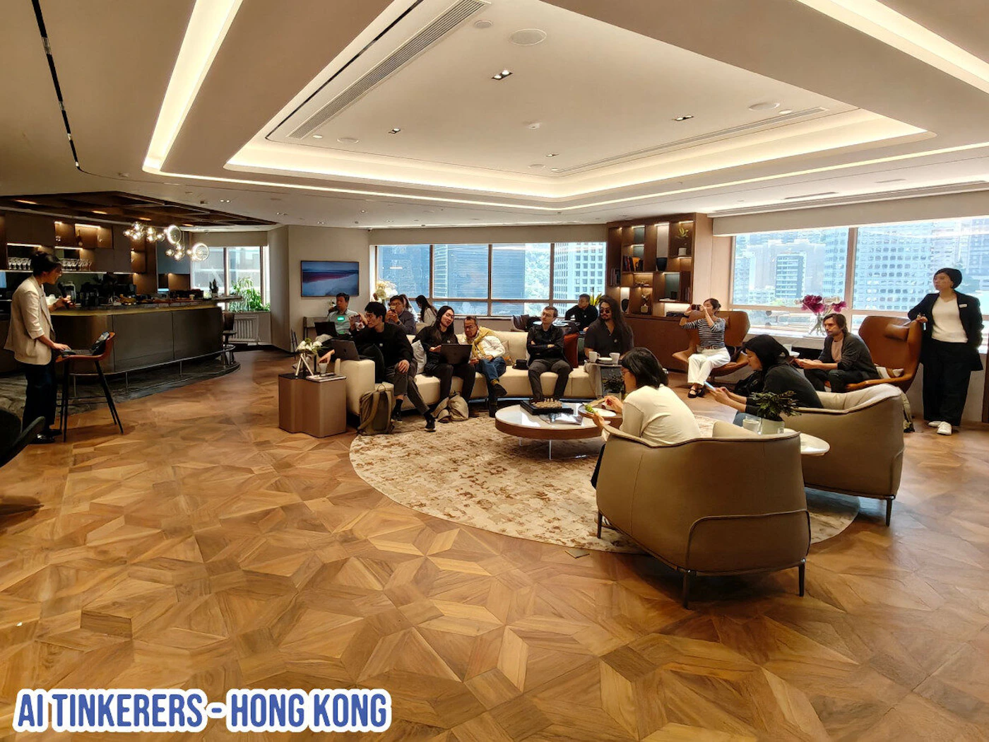 (Photo) The image shows a group of people gathered in a modern office or event space, possibly attending a presentation or meeting. There is a speaker standing in front of the group. Text: AI TINKERERS - HONG KONG group of people | indoor | presentation, modern office space, large windows | candid | Colors: #D2B48C, #000000, #FFFFFF Note: The image depicts a real-world scene with people, furniture, and architectural elements, making it a photograph. The presence of recognizable objects and people suggests it's not an illustration or abstract art.