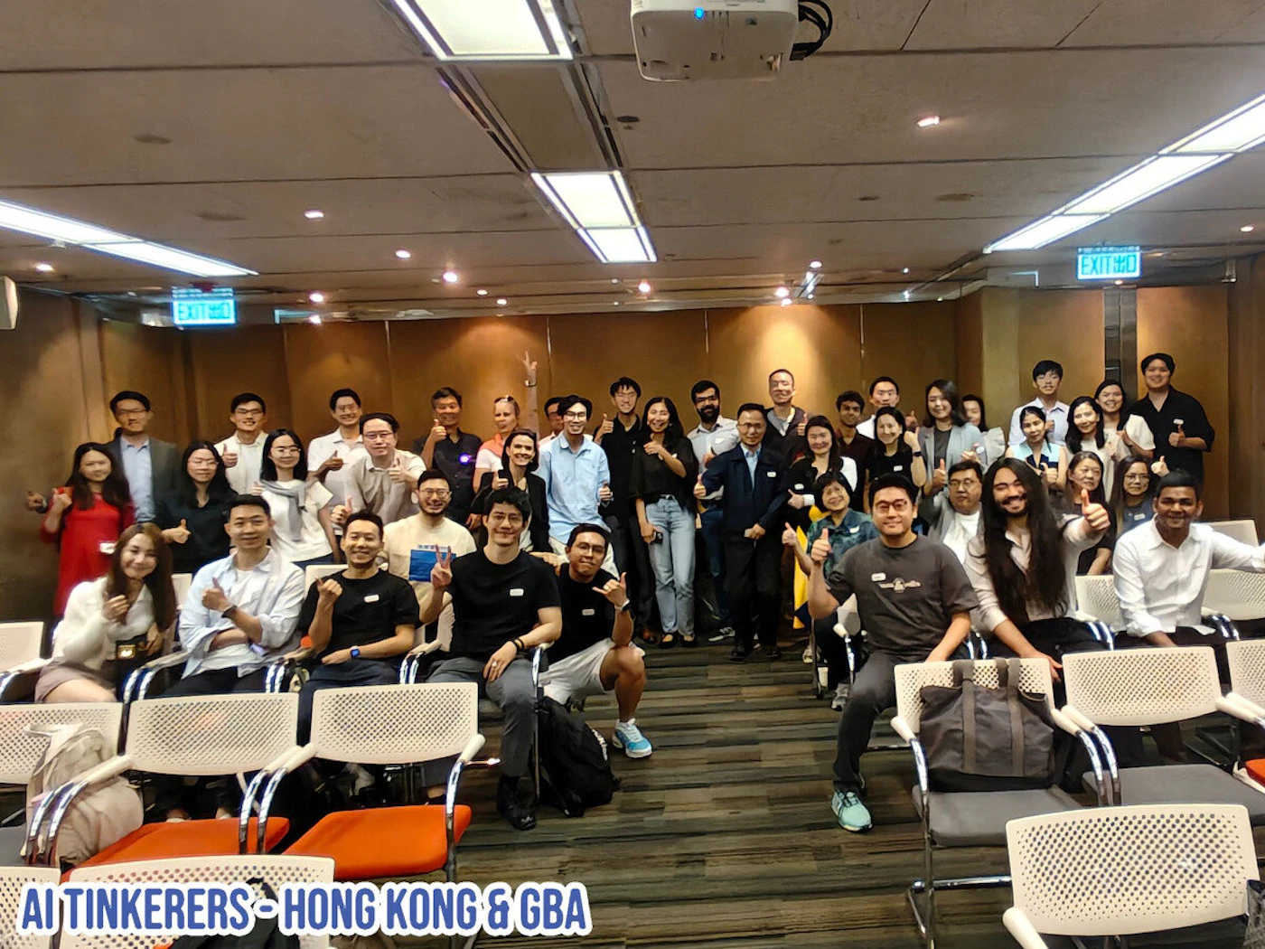 (Photo) A group of people are posing for a photo in what appears to be a conference room. They are giving thumbs up. Text: AI TINKERERS - HONG KONG & GBA group of people | indoor | multiple people, thumbs up gesture | candid | Colors: #FFFFFF, #000000, #D2B48C Note: The image is a real-world scene captured by a camera, making it a photograph. It shows a group of people in a room.