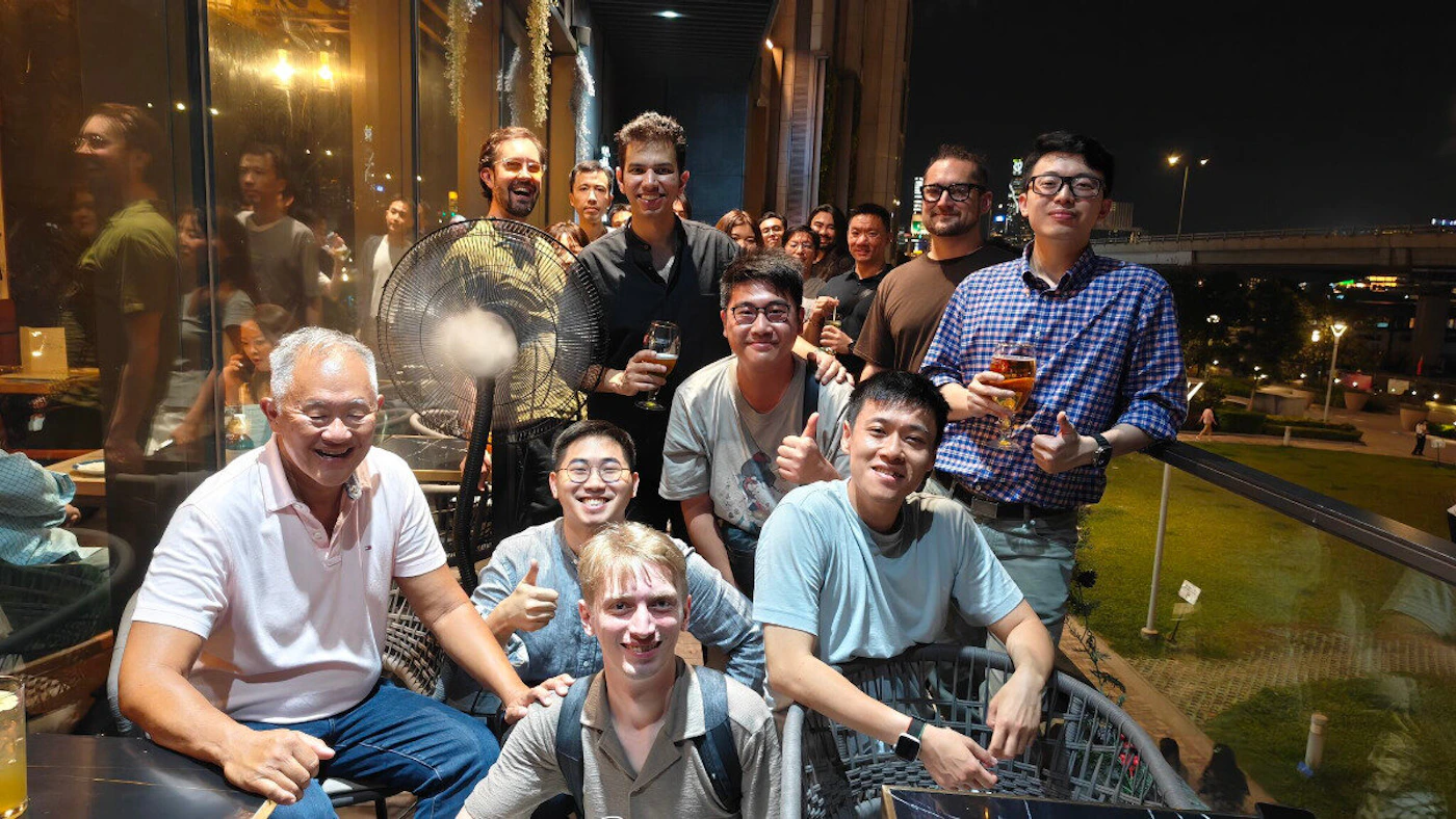 (Photo) A diverse group of people, predominantly men, are gathered on an outdoor patio or balcony at night, smiling and holding drinks. The background features city lights, buildings, and some greenery. group of people, social gathering | outdoor patio/balcony, urban night | multiple people, some holding drinks, a fan, city lights in background, reflections on glass | candid/group portrait, night photography | Colors: #1A1A1A, #A08040, #E0B49A, #4060A0, #E0C0C0 Note: The image is a direct capture of a real-world scene featuring multiple individuals in a social setting, which is characteristic of a photograph.