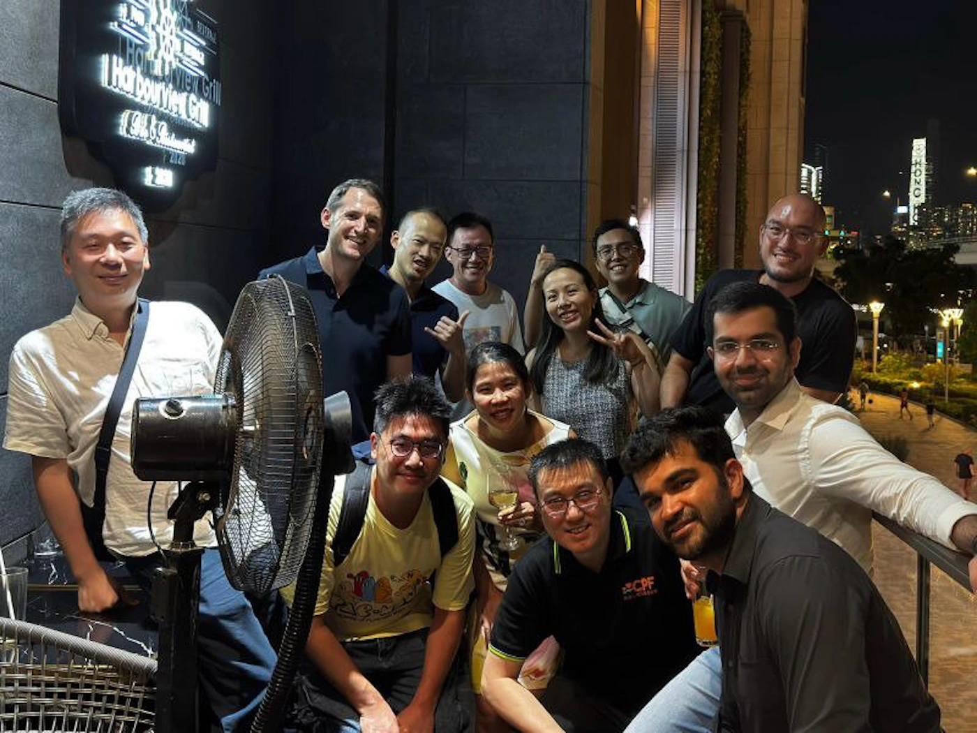 (Photo) A group of about twelve adults, appearing to be friends or colleagues, are posing for a photo on an outdoor patio at night, with a glowing restaurant sign on a dark wall behind them and city lights in the distant background. Text: THE [unreadable symbol] 1 Harbourview Grill [unreadable] 2128 group of people | outdoor patio at night, urban environment | multiple adults, city lights in background, glowing restaurant sign, large standing fan in foreground | candid group photo | Colors: #2F2F2F, #C09B7C, #F0F0F0, #FFD700 Note: The image is a realistic capture of a moment, featuring a group of people in a real-world setting with natural lighting, characteristic of a photograph.