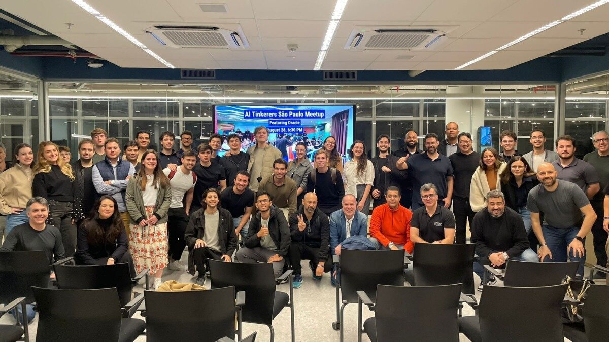 A group of approximately 40-50 people, appearing to be participants or attendees of an event, are posing for a photo indoors. A large screen in the background displays event details for an 'AI Tinkerers São Paulo Meetup'.. Text: AI Tinkerers São Paulo Meetup
Featuring Oracle
August 28, 6:30 PM
