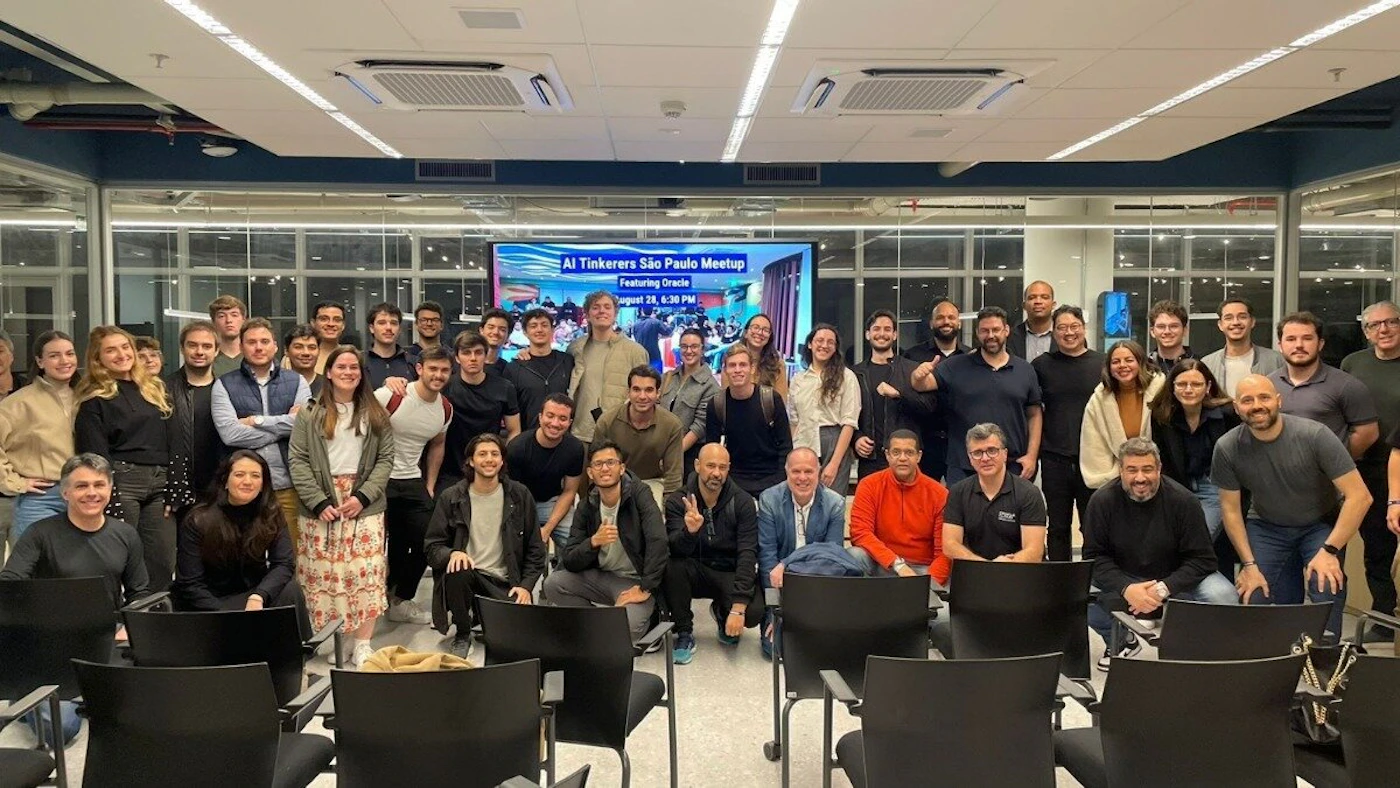 (Photo) A large group of approximately 50-60 adults, both men and women, are gathered in an indoor event space, posing for a group photograph. A large screen in the background displays information for an 'AI Tinkerers São Paulo Meetup'. Text: AI Tinkerers São Paulo Meetup Featuring Oracle August 28, 6:30 PM group of people at an event | indoor event space or office | approximately 50-60 adults, a large screen displaying event details, modern interior design, many people smiling | group portrait, posed | Colors: #333333, #F0F0F0, #A0A0A0, #4682B4, #D3D3D3 Note: The image is a direct capture of a real-world scene, featuring a large group of people posing together at an event, which is characteristic of a photograph.