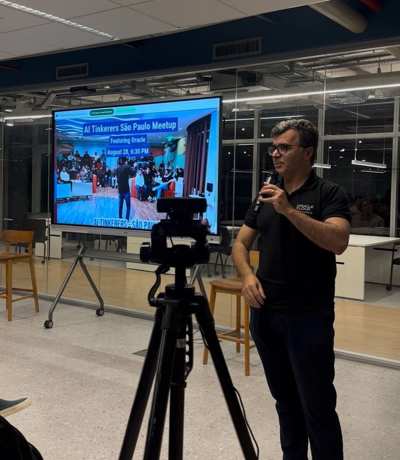 (Photo) The image captures a man speaking into a microphone during an indoor event, with a large screen behind him displaying a presentation slide for an "AI Tinkers São Paulo Meetup." A camera on a tripod is positioned in the foreground, suggesting the event is being recorded or streamed. Text: AI Tinkers São Paulo Meetup Featuring Oracle August 28, 6:30 PM AI TINKERERS - SÃO PAULO ORACLE Cloud Infrastructure A speaker at a professional event, a presentation screen, and event recording equipment. | Indoor, modern office or event space with glass walls and contemporary furniture. | A man in a black polo shirt with an 'ORACLE Cloud Infrastructure' logo speaking into a microphone, a large screen displaying event details and an image of an audience, and a camera on a tripod. | Candid, event photography. | Colors: #1A1A1A, #E0E0E0, #0066CC, #A06030 Note: The image is a realistic capture of a scene featuring people, objects, and an environment, characteristic of a photograph. It shows an actual event in progress rather than a stylized illustration, logo, or handwritten note.