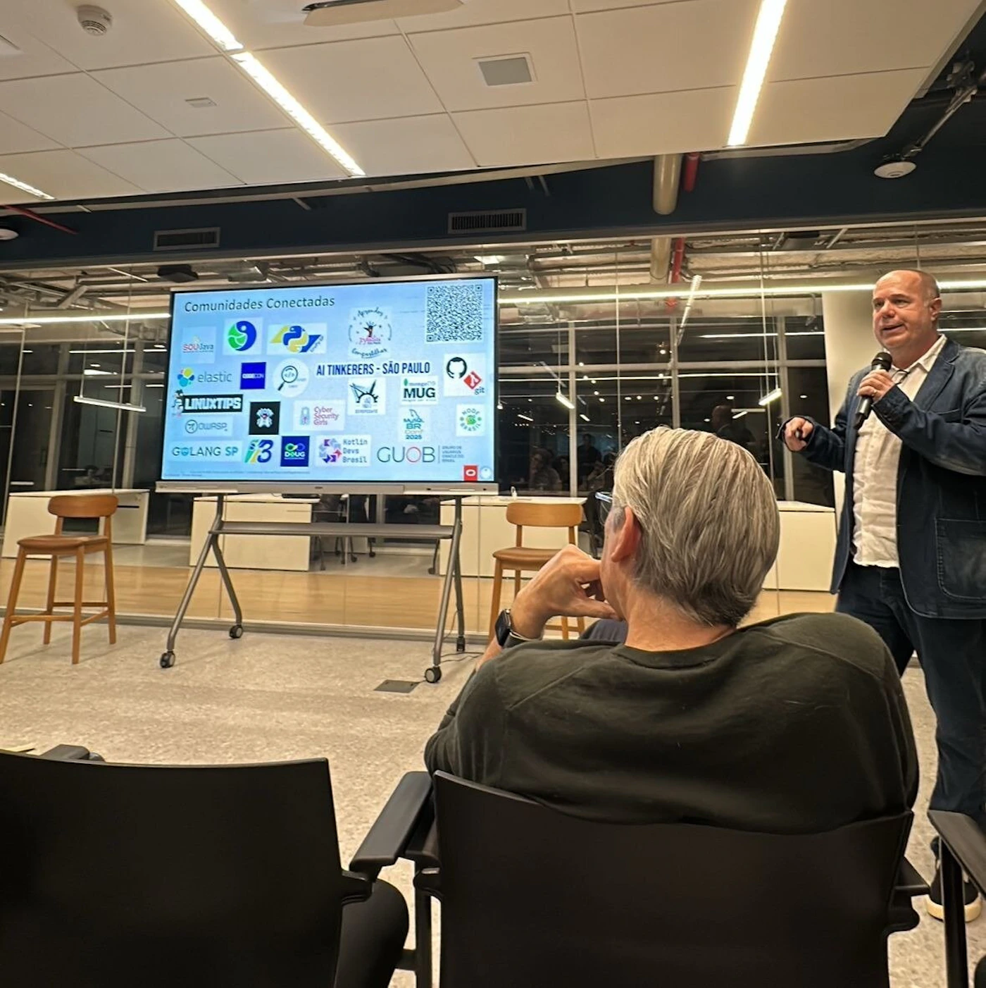 (Photo) A photo captures an indoor presentation setting where a man is speaking into a microphone, standing next to a large screen. The screen displays a title 'Comunidades Conectadas' along with numerous logos and names of technology communities. An audience member is visible from behind in the foreground. Text: Comunidades Conectadas elastic LINUXTIPS GOLANG SP AI TINKERERS - SÃO PAULO MUG Cyber Security Skills Kotlin Brasil GUOB [unreadable text on various logos] A presentation or conference event | Indoor, modern office or event venue | A speaker holding a microphone, a large digital screen displaying various community logos and text, an audience member in the foreground. | Candid, event photography | Colors: #4A4A4A, #FFFFFF, #8C8C8C, #2E7D32, #1976D2 Note: The image is a realistic photographic capture of a real-world event or presentation, featuring people and a setting, rather than a stylized illustration, logo, banner, or handwriting.