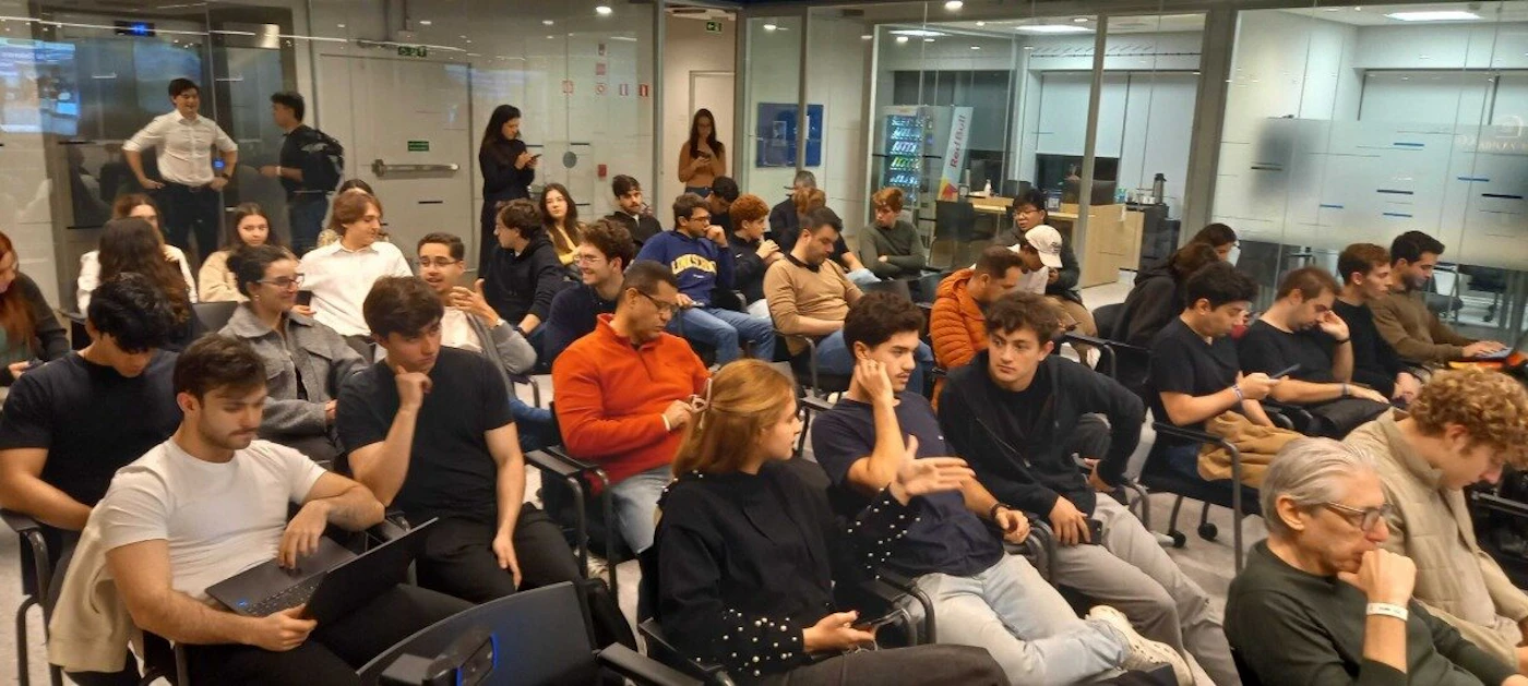 (Photo) The image captures a diverse group of people, primarily young adults, gathered in a modern indoor setting that resembles a classroom or meeting space. Attendees are seated in rows, some engaged in conversation, others looking forward, and a few using laptops or mobile phones. group of people, audience, meeting | indoor, modern office or classroom | multiple people (mostly young adults), glass partitions, some individuals using laptops or phones, casual attire | candid, wide shot | Colors: #505050, #A0A0A0, #202020, #D0D0D0, #806040 Note: This image is classified as a 'photo' because it is a realistic capture of a real-world scene, depicting people and an indoor environment with natural lighting and perspective, rather than an illustration, logo, banner, or handwriting.