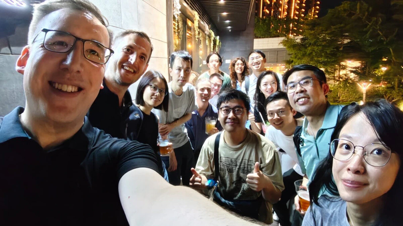 (Photo) A group selfie captures a diverse group of approximately 15-18 individuals smiling and holding drinks in an urban outdoor setting at night, with city lights and foliage visible in the background. A group of people | Outdoor, urban, night, possibly a bar or restaurant patio | Multiple people (diverse group), smiling, some holding drinks, city lights in background, selfie perspective. | Candid, group selfie | Colors: #1A1A1A, #E0B49F, #F5B041, #4CAF50, #FFD700 Note: The image is a direct capture of a real-world scene featuring people, characteristic of a photograph rather than an illustration, logo, or text-based content.