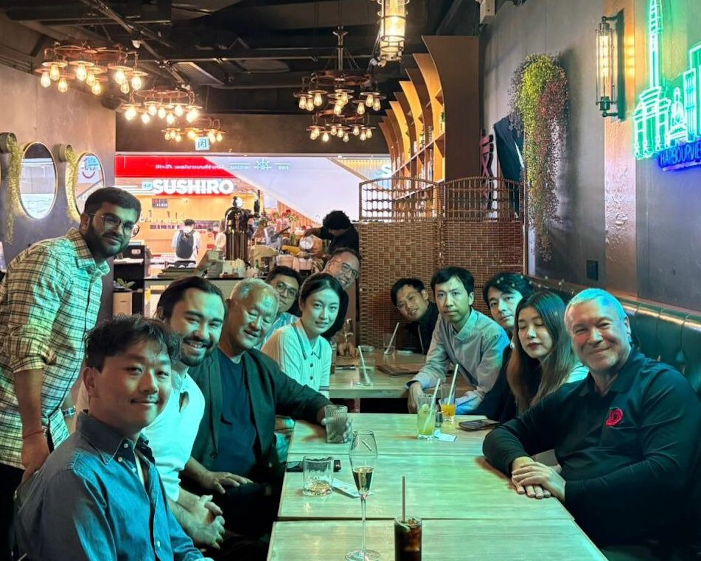 (Photo) A group portrait of about a dozen people seated together at a large wooden table inside a dimly lit restaurant or bar. Text: SUSHIRO HARBOURVIE A diverse group of adults | Indoor restaurant or bar, characterized by industrial lighting and dark walls. | Group portrait, low-light environment, visible signage for 'SUSHIRO' and a neon sign partially reading 'HARBOURVIE' in the background. | Group portrait, candid, low light photography | Colors: #3D3D3D, #C87A3E, #70A39B, #F5F5F5 Note: This image is a captured moment of real people gathered in a real indoor setting, making it a standard photograph.