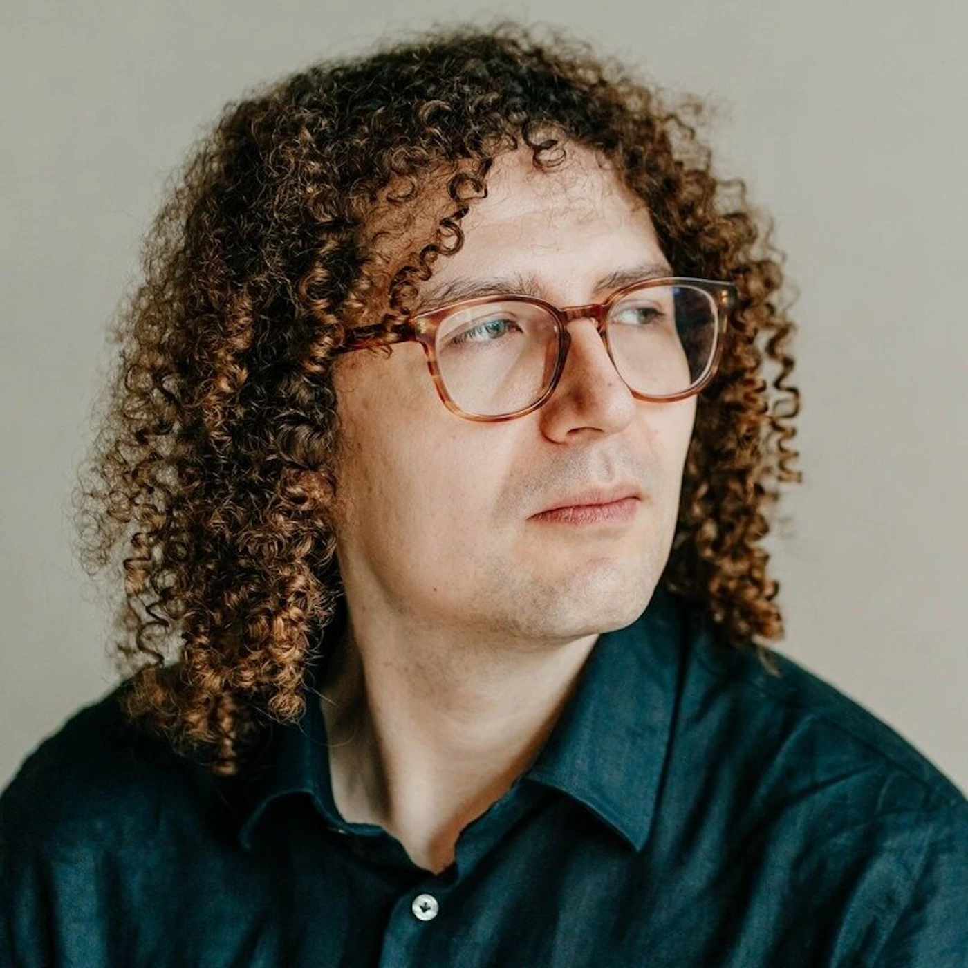 (Photo) A portrait of a man with voluminous curly brown hair and glasses, looking thoughtfully to the side against a neutral background. person | indoor | curly hair, glasses, neutral background | portrait | Colors: #d8d1c6, #1a2a33, #7b5038 Note: The image is a clear, high-resolution photograph of a human subject, characteristic of a professional portrait or headshot.