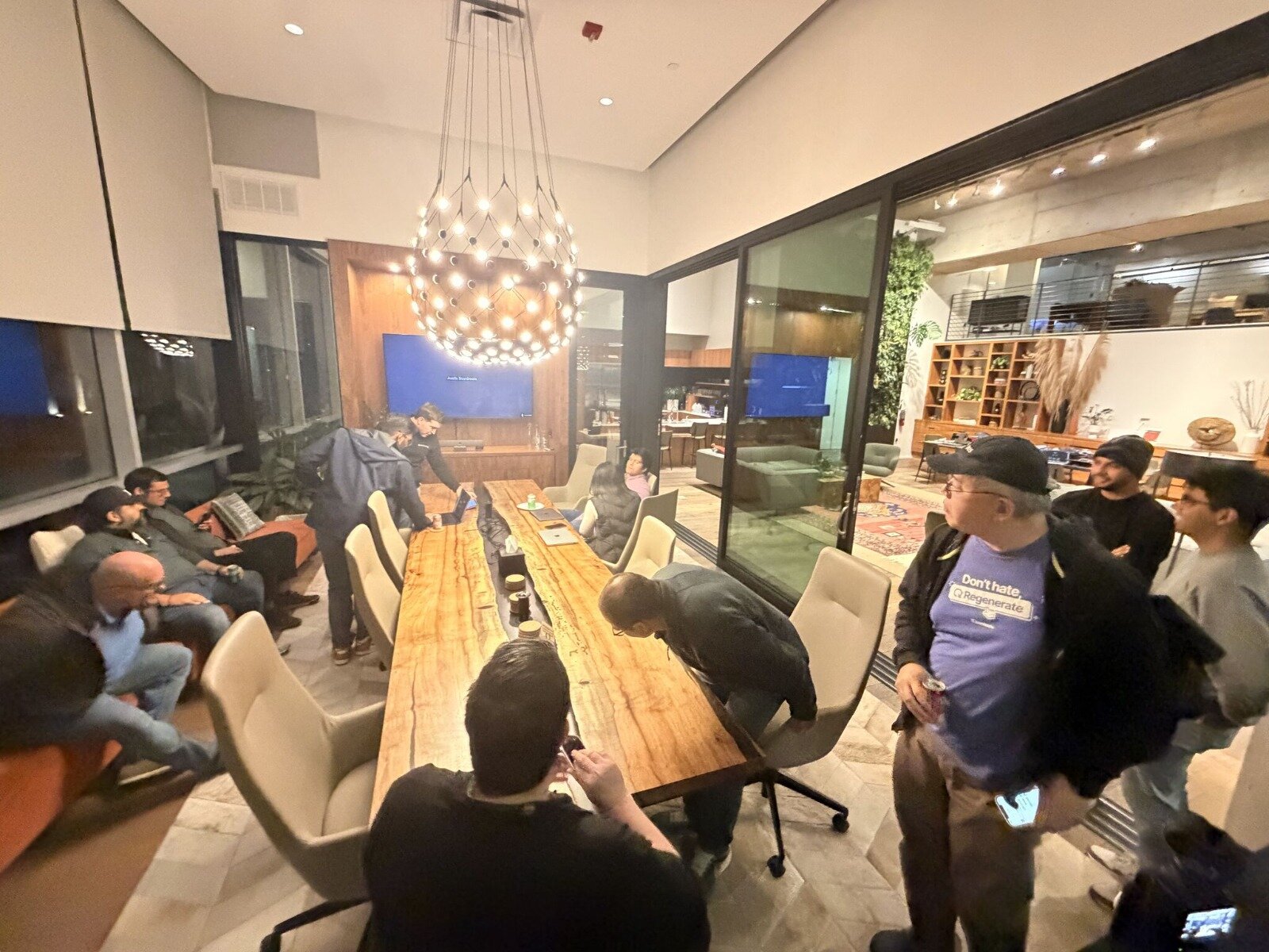 A candid photograph captures a group of approximately ten people gathered around a long, rustic wooden conference table in a modern, well-lit indoor meeting space.. Text: Don't hate, Regenerate.