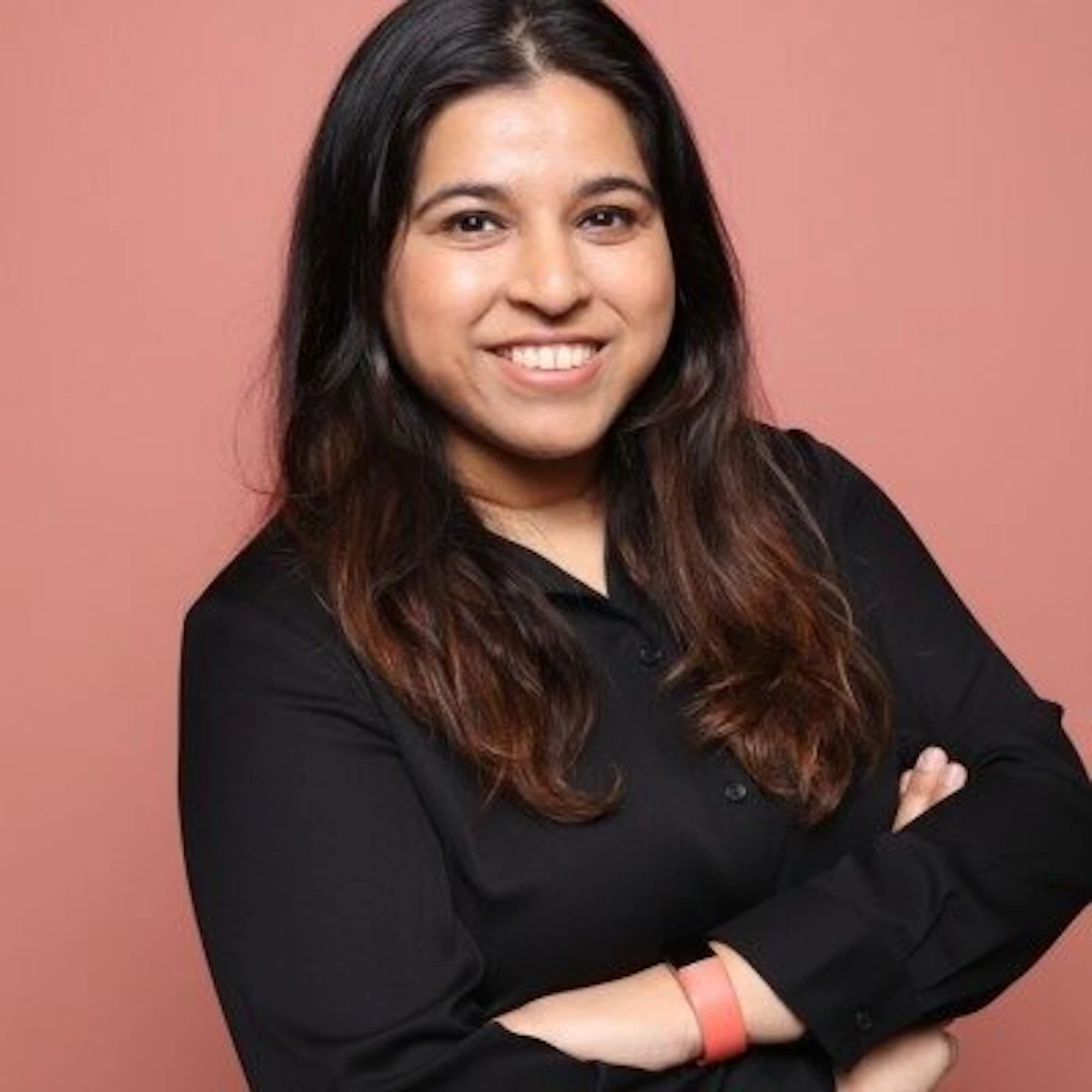 (Photo) A studio portrait of a woman with long dark hair, wearing a black shirt and smiling, set against a solid pink background. person | indoor | smiling woman, solid pink background, arms crossed | portrait | Colors: #d18a80, #1a1617, #4a312d Note: This is a real-life photograph of a person, characteristic of a professional headshot or studio portrait.