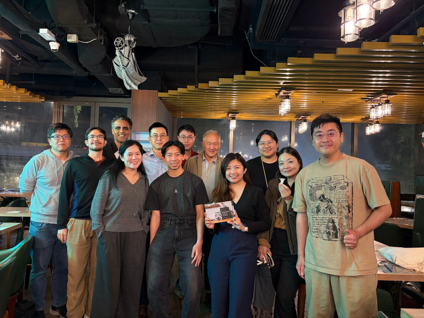 (Photo) A group of twelve people posing for a group photo in what appears to be a restaurant or social venue with warm lighting. Text: THE CAT MIMIC group of people | indoor restaurant | group posing, indoor lighting, casual attire, wooden ceiling slats | group portrait | Colors: #c5b497, #222222, #4a5d7a, #e0e0e0 Note: The image is a real-life photograph capturing a group of individuals in a physical indoor environment.