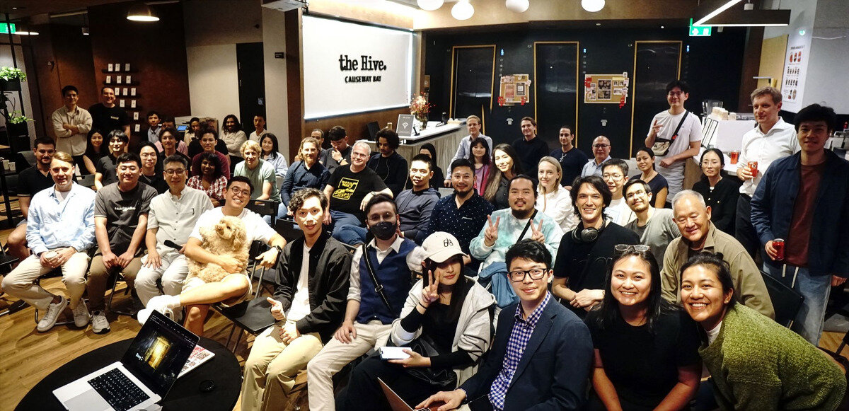 A large group of people are gathered for a group photo in an indoor event space, with one man in the front row holding a small dog.. Text: the Hive. CAUSEWAY BAY