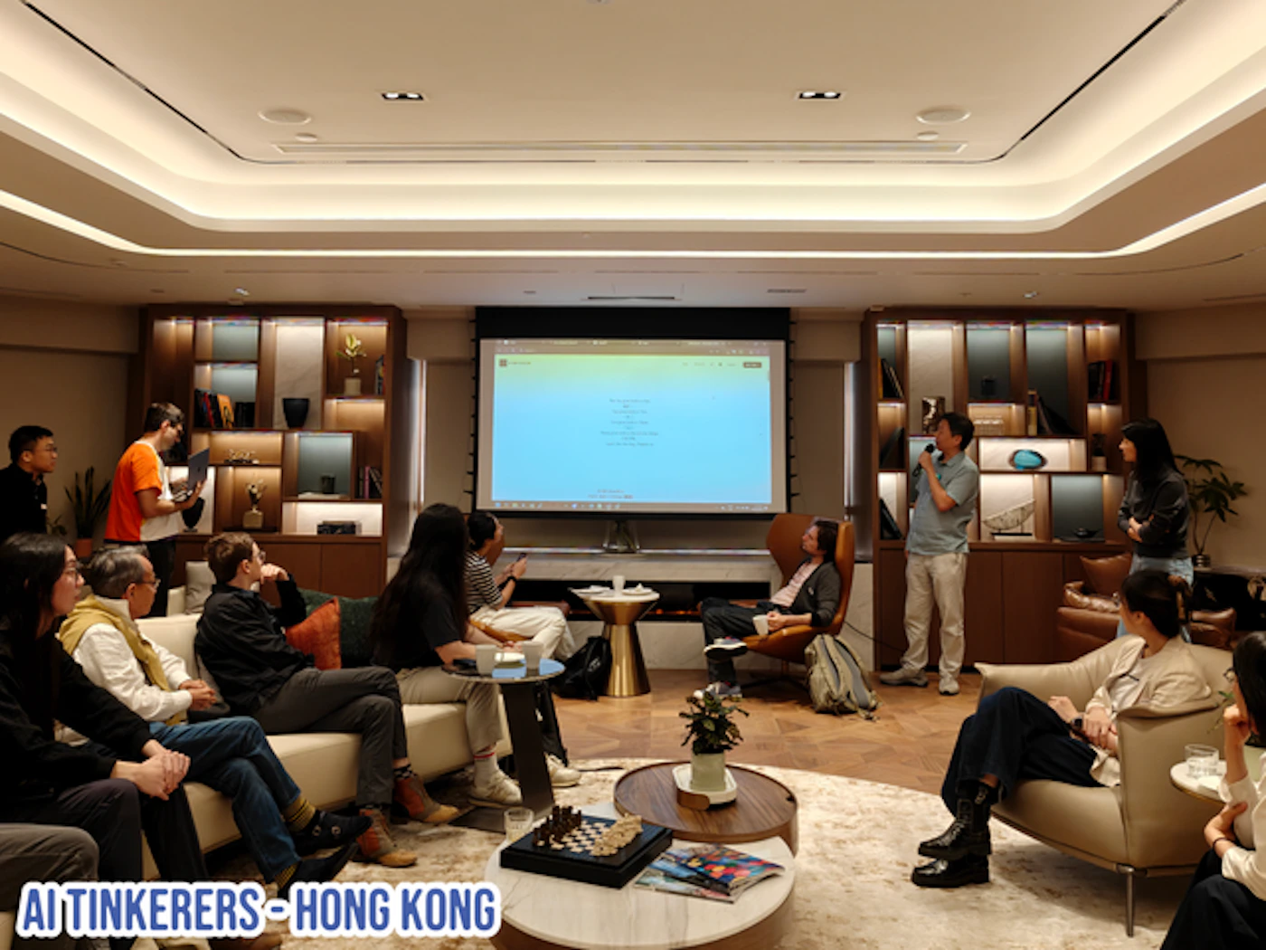 (Photo) A photo shows a group of people gathered in a room, watching a presentation on a projector screen. There is a presenter speaking to the audience. Text: AI TINKERERS - HONG KONG people | indoor | group of people, projector screen, presentation | candid | Colors: #D3CEC4, #282424, #FFFFFF Note: The image depicts a real-life scene with people and objects, suggesting it is a photograph rather than an illustration or other image type.