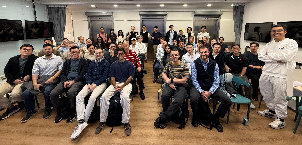 A large group of approximately fifty people, mostly men, are gathered indoors in a brightly lit room, posing for a group photograph.